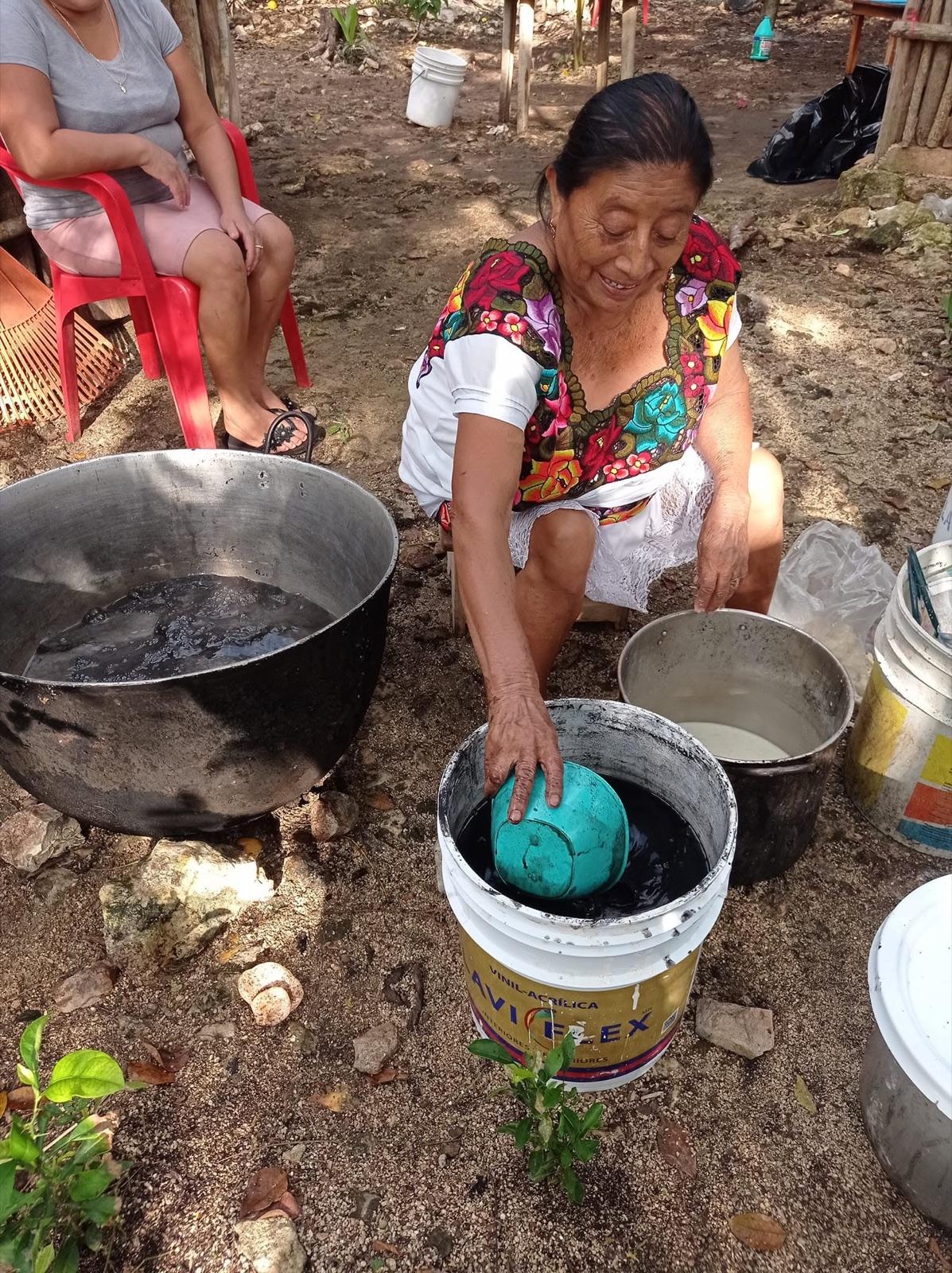 Mayan grandmother in a colorful embroidered huipil crouching to work over metal pots and a bucket at a jungle homestead