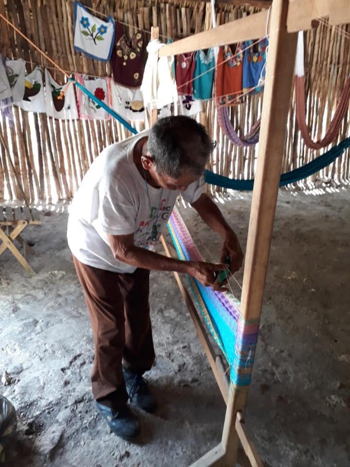 A man weaving colorful threads on a wooden loom inside a palapa hung with embroidered textiles