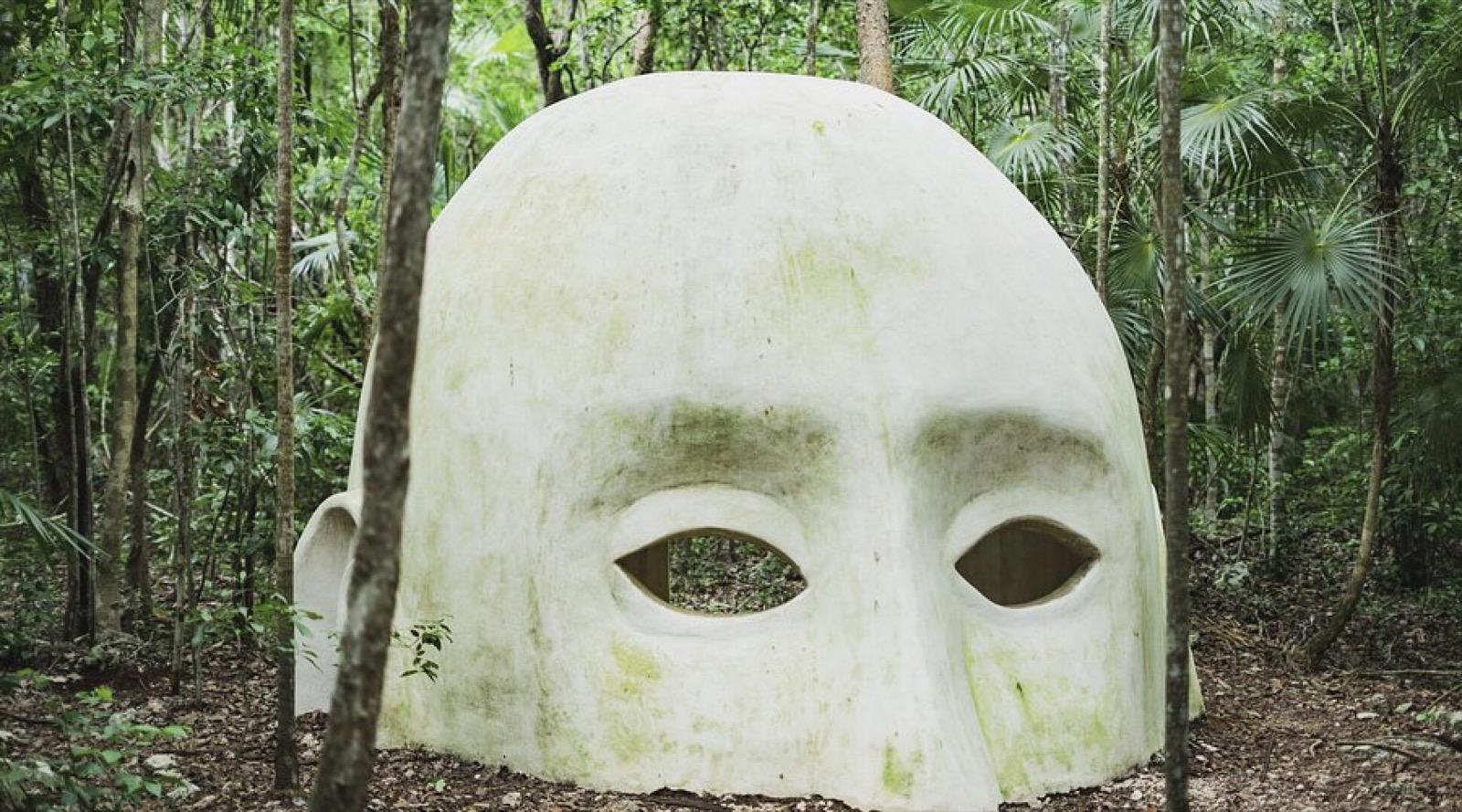 A giant pale face sculpture with hollow eyes standing among the jungle trees at Holistika, Tulum.