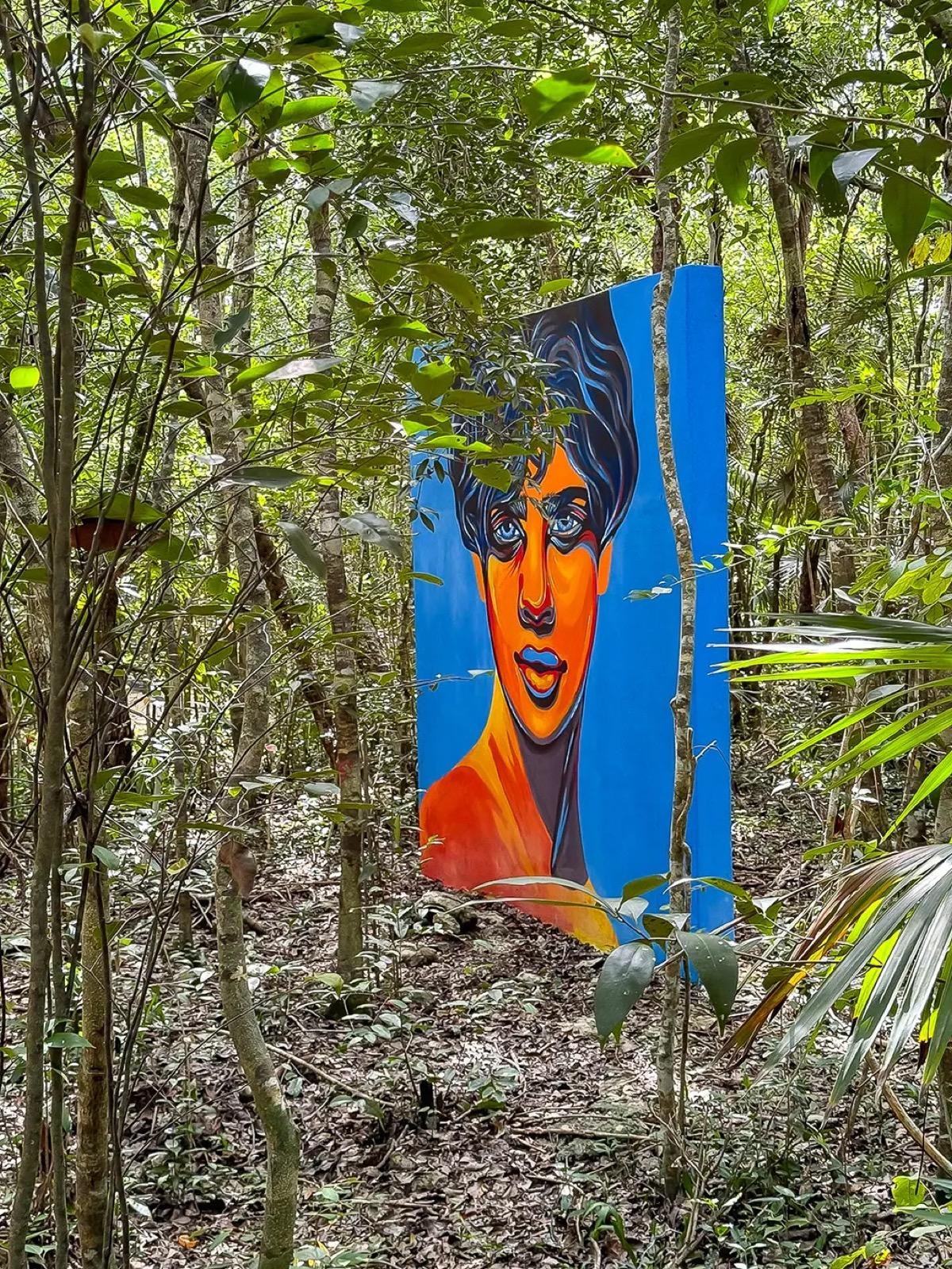 A colorful mural of a face painted on a tall blue panel set among the trees of the Tulum jungle.