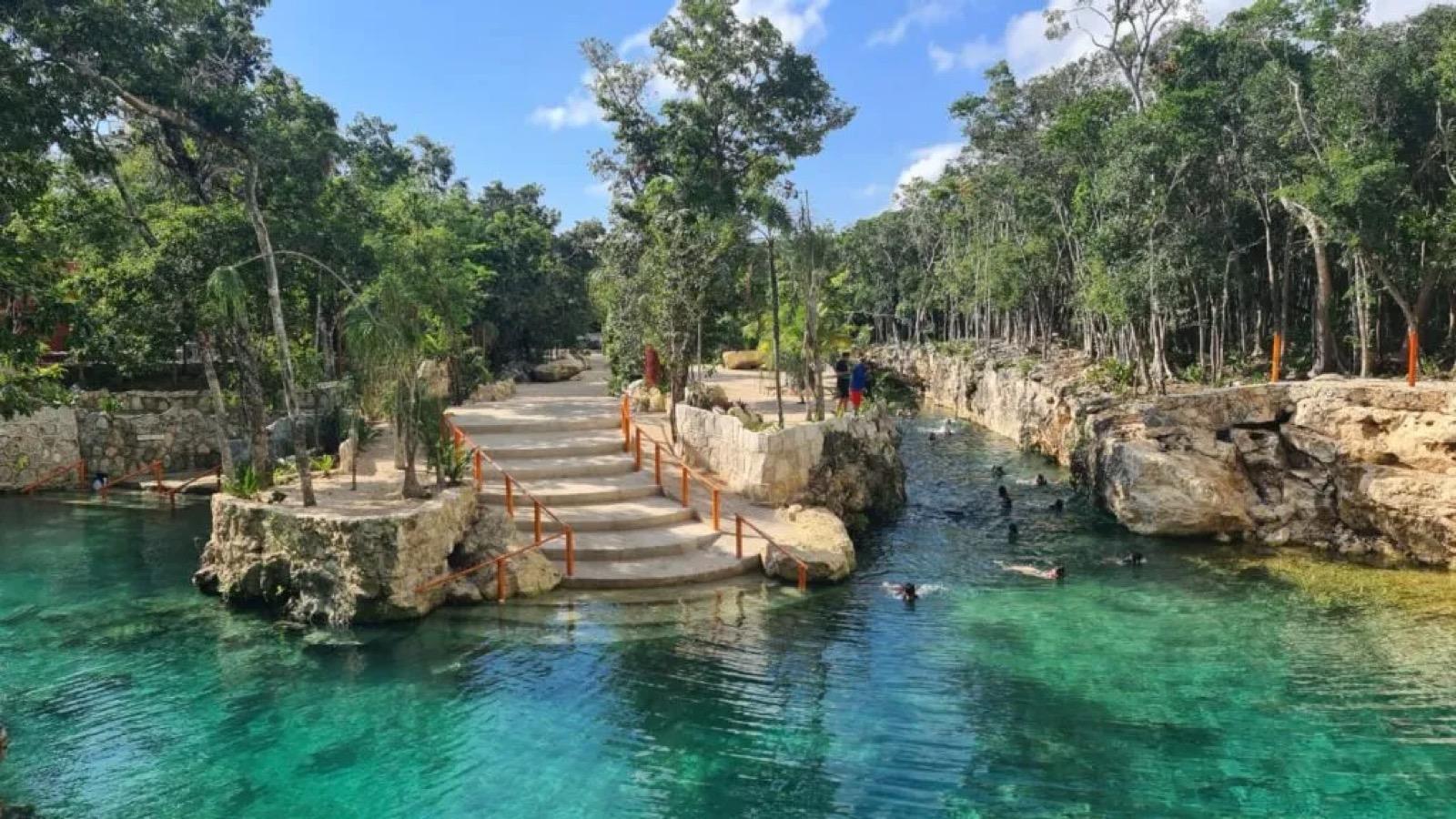 Un cenote a cielo abierto de agua turquesa con escalones de piedra y una pasarela de madera entre la selva.