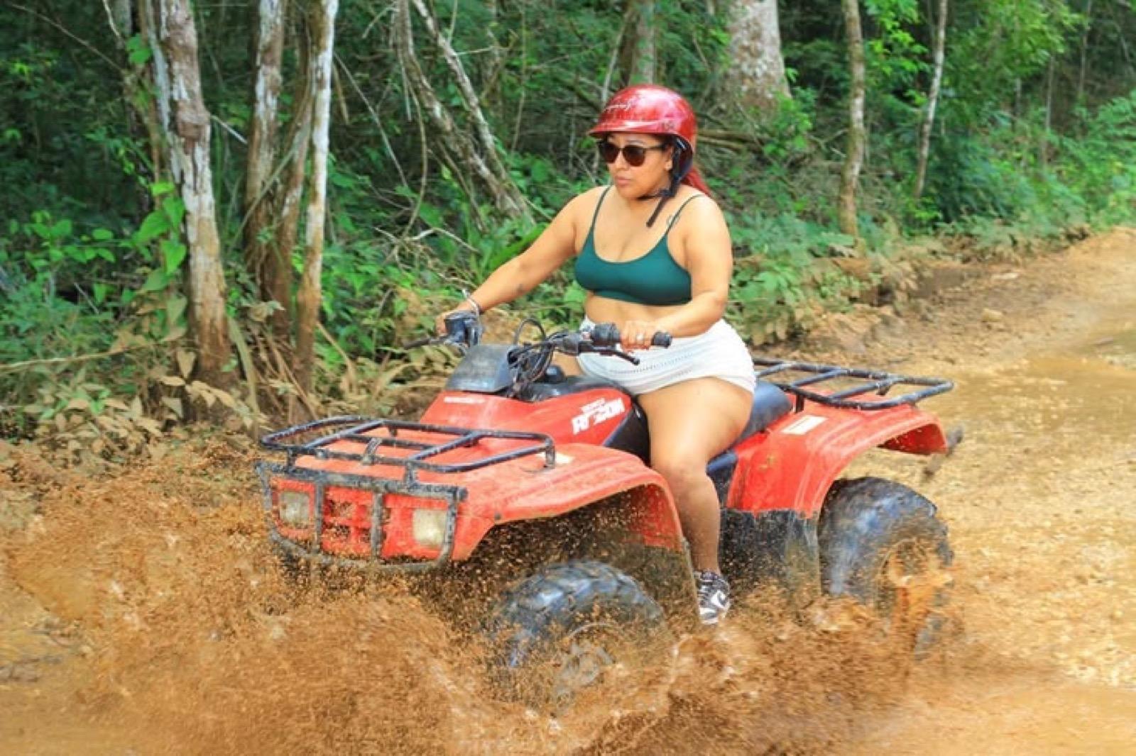Una conductora salpicando lodo en una cuatrimoto roja por un camino de selva entre los árboles.