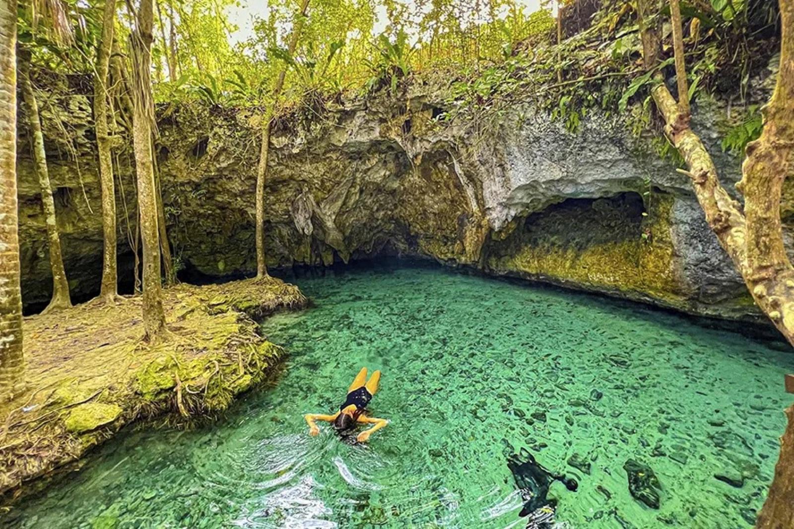 Swimmer floating in a turquoise open-air jungle cenote framed by rock overhangs and greenery