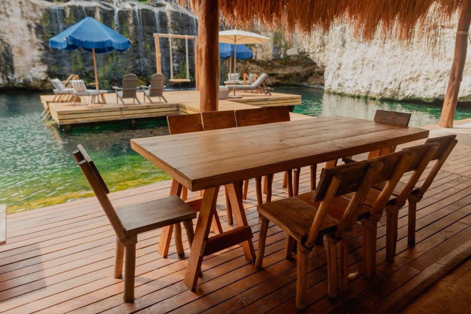 A long wooden dining table under a palapa on the deck, with umbrellas and the cenote behind.