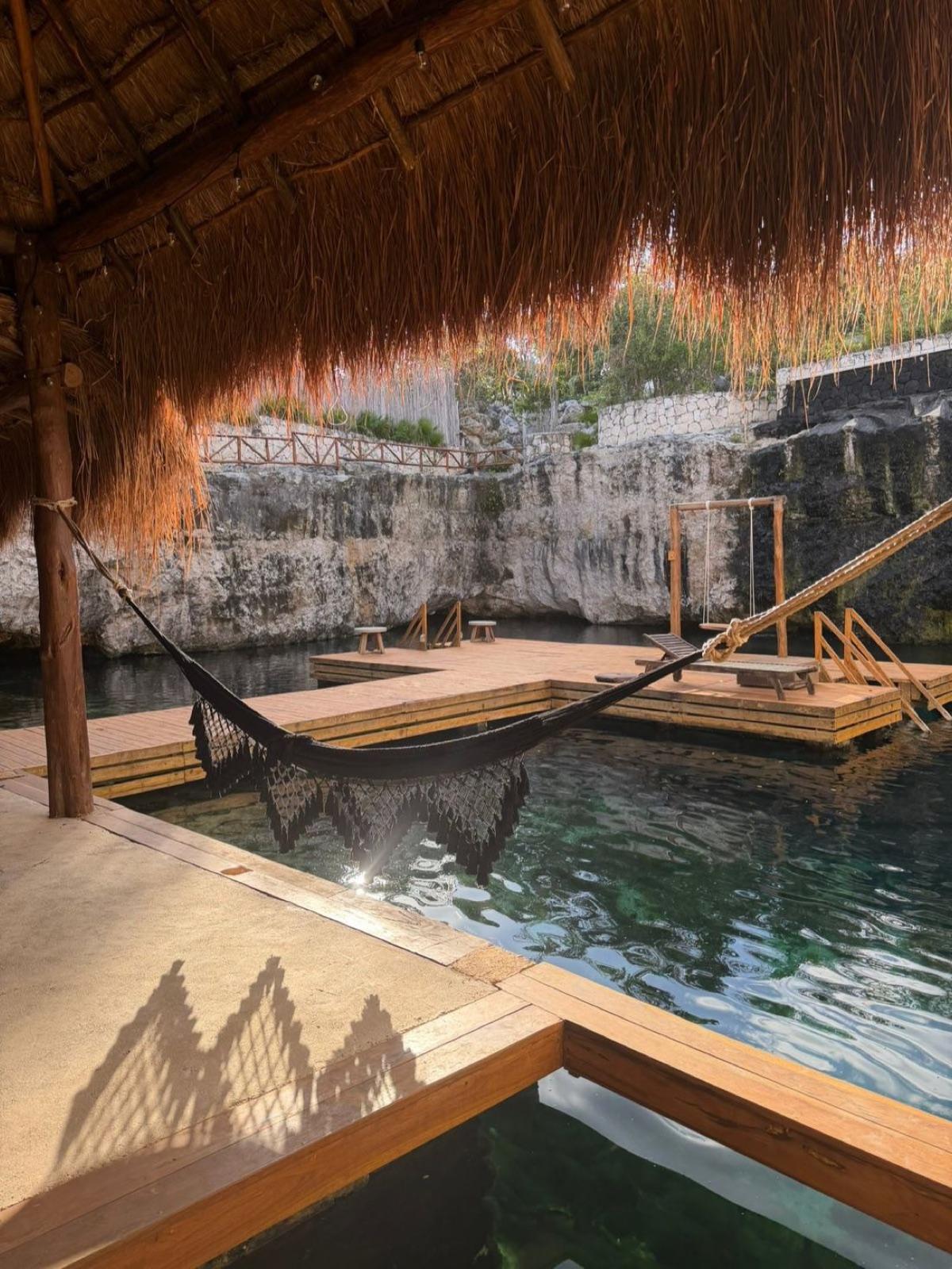 A hammock strung under a thatched palapa over the cenote water, deck and stone walls around.