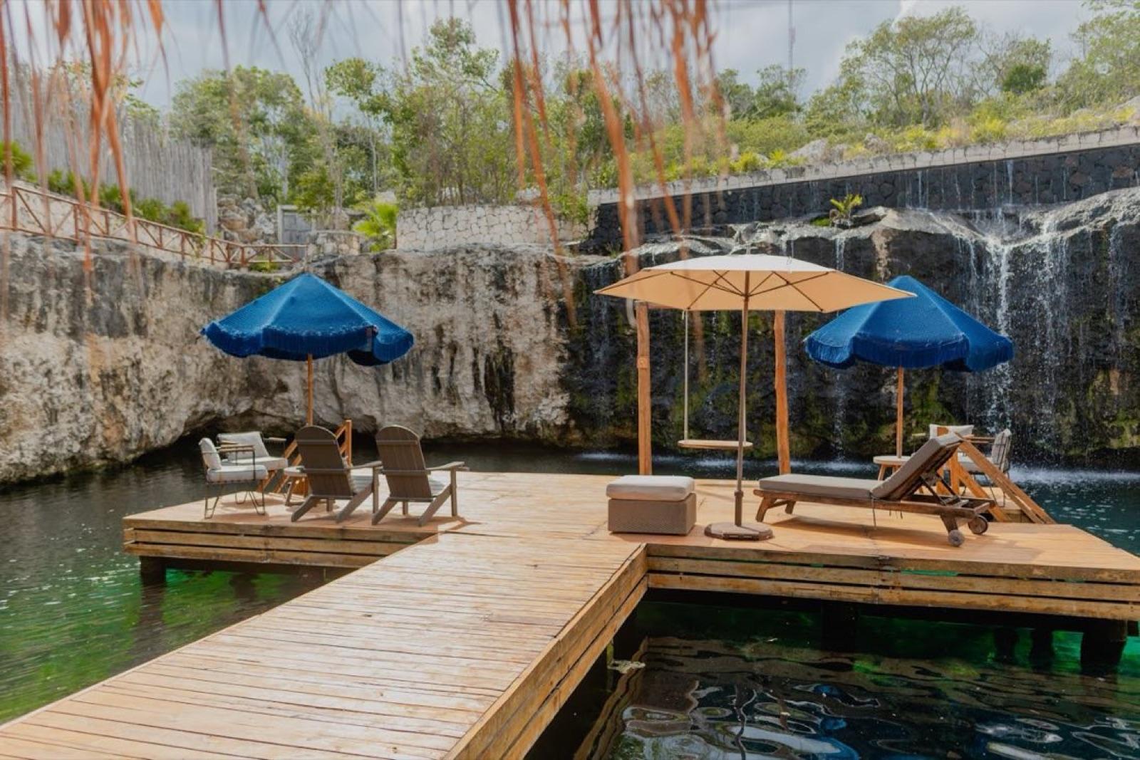 A private cenote with a wooden sun deck, blue umbrellas and loungers over clear turquoise water, ringed by stone walls.