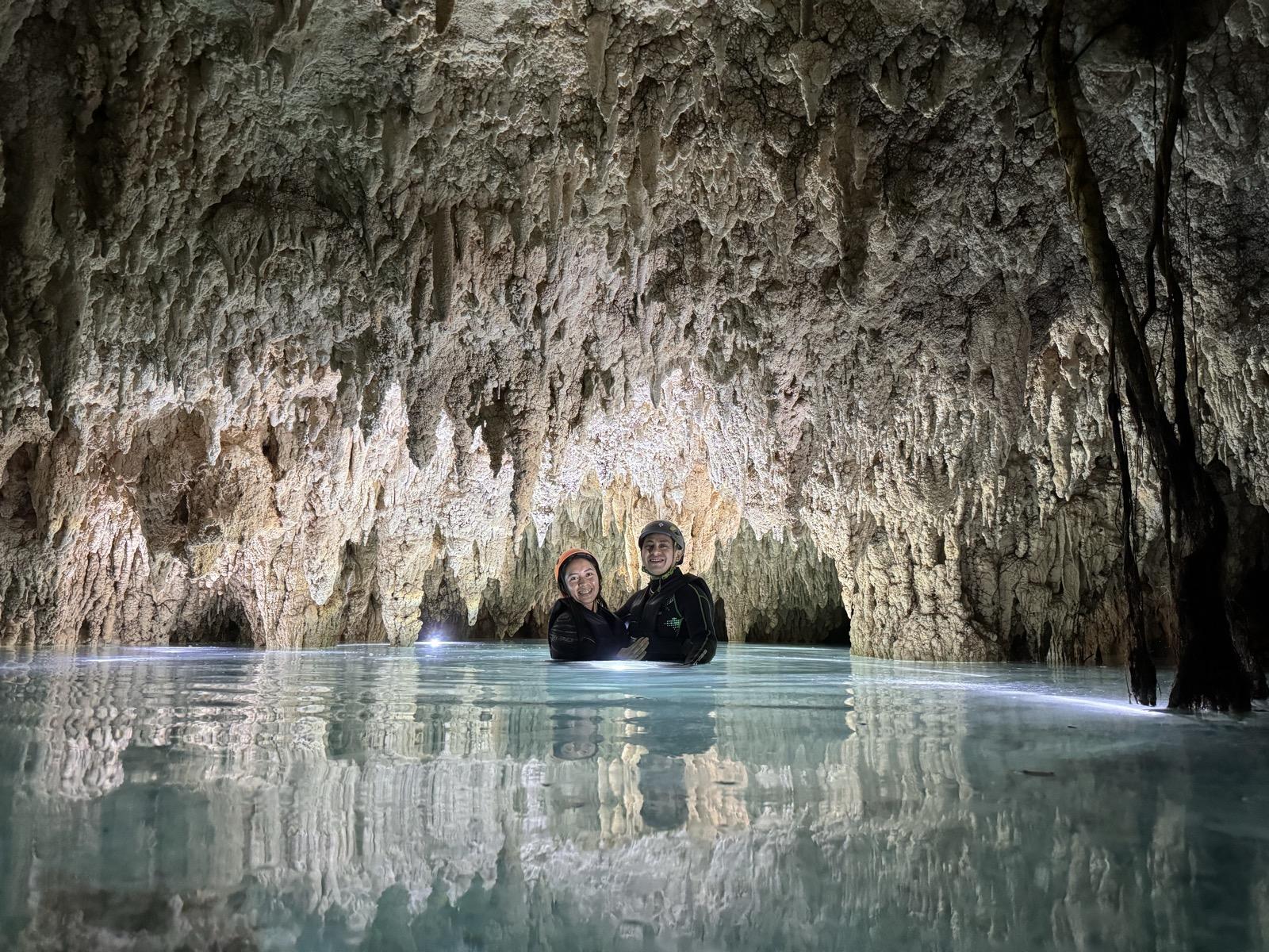 Inside a cave cenote, two people wade through calm turquoise water beneath a stalactite ceiling.