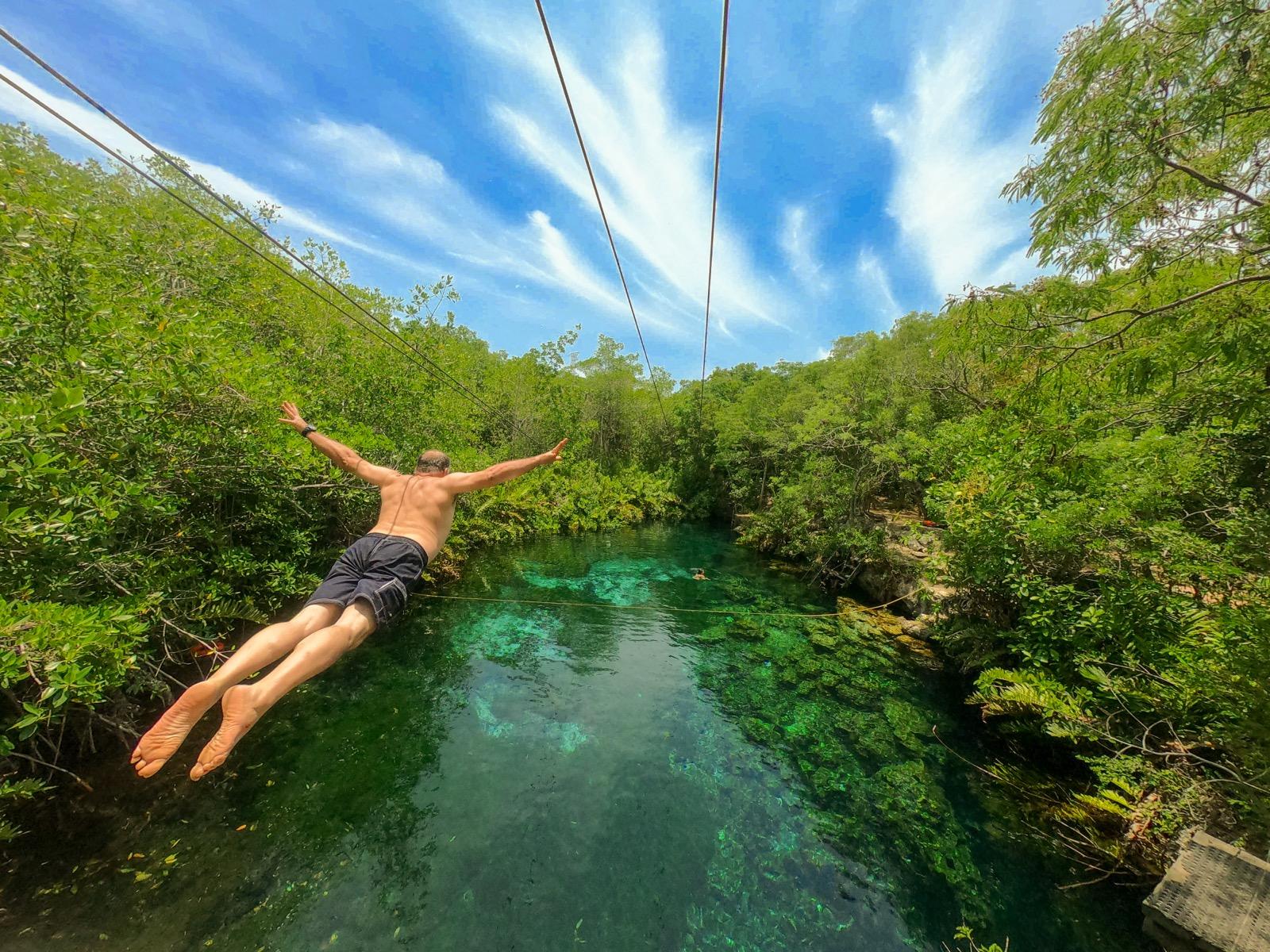 A swimmer leaps arms-open into a turquoise open-air cenote ringed by green jungle.