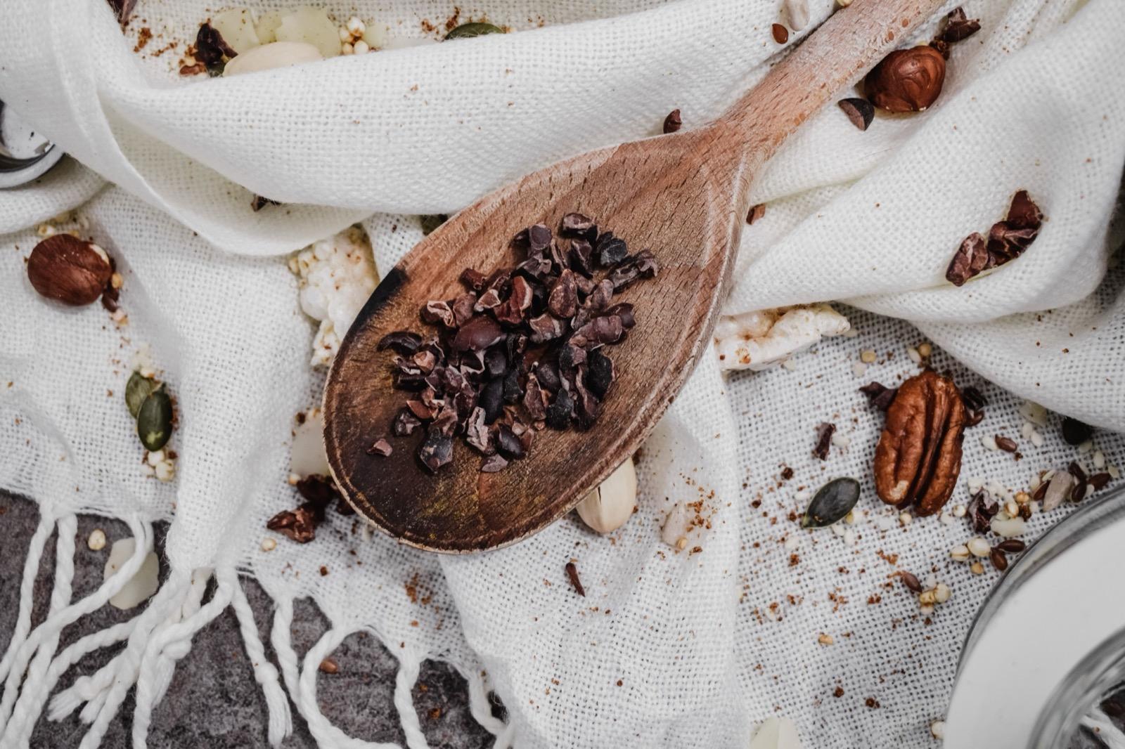 A wooden spoon of cacao nibs and beans arranged on a white cloth, seen from above.