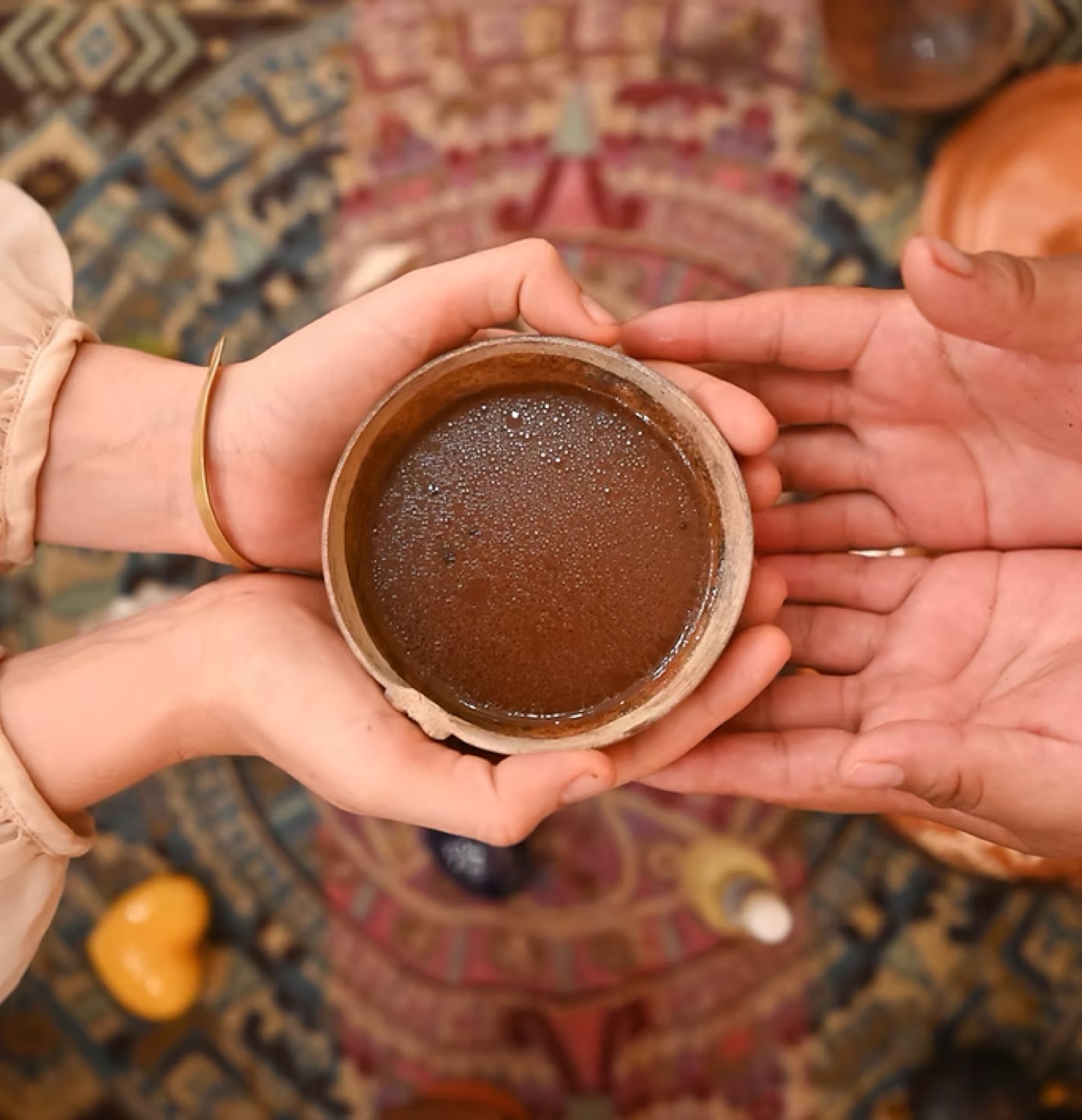 Hands cradling a rustic clay cup filled with warm ceremonial cacao over a patterned textile altar.