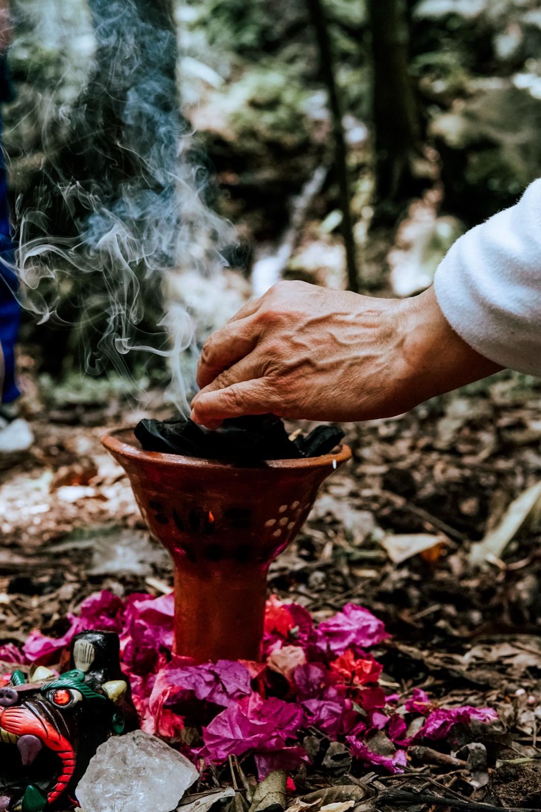 Hands making an offering over a clay cup as copal smoke rises, flowers arranged around it in the jungle.
