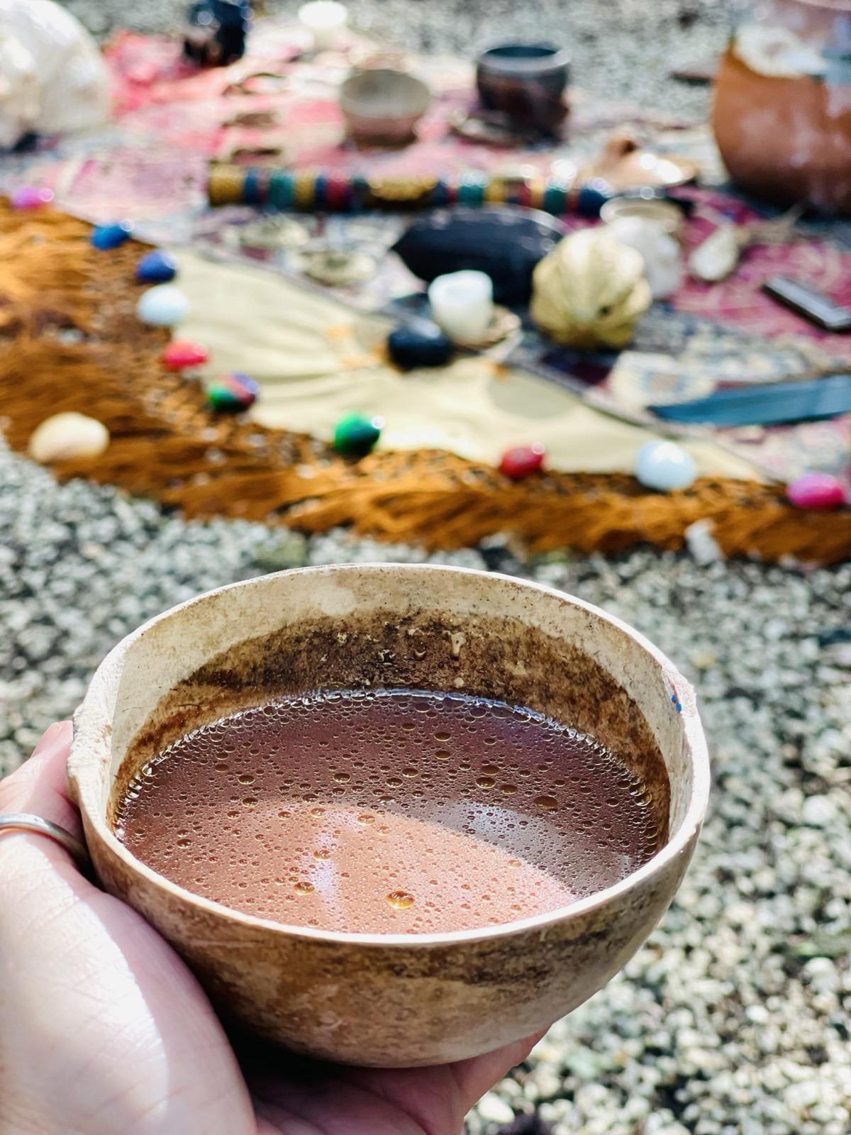 A hand holding a cup of ceremonial cacao above an altar laid with crystals and offerings.