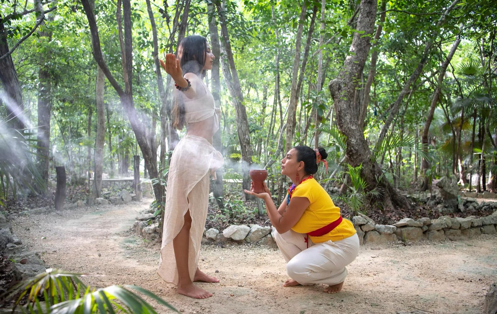 Two women dressed in white during an outdoor cacao ceremony in a sunlit jungle clearing, one standing with arms raised and the other kneeling with a cup as mist drifts among the trees.