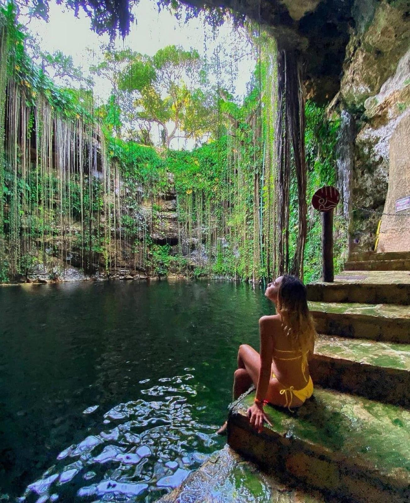 El cenote Ik Kil de aguas turquesa y raíces colgantes que caen desde el borde, con una visitante descansando en los escalones de piedra