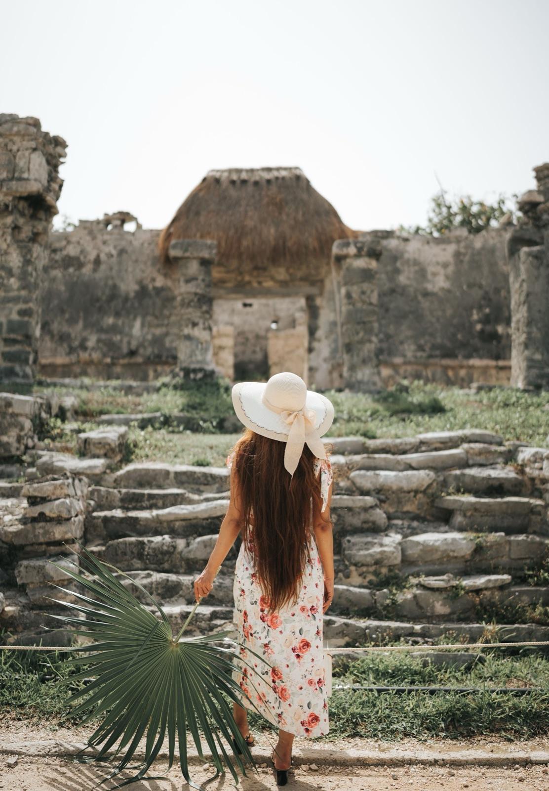 Una visitante con sombrero y vestido floreado camina entre las ruinas de piedra de una antigua ciudad maya.