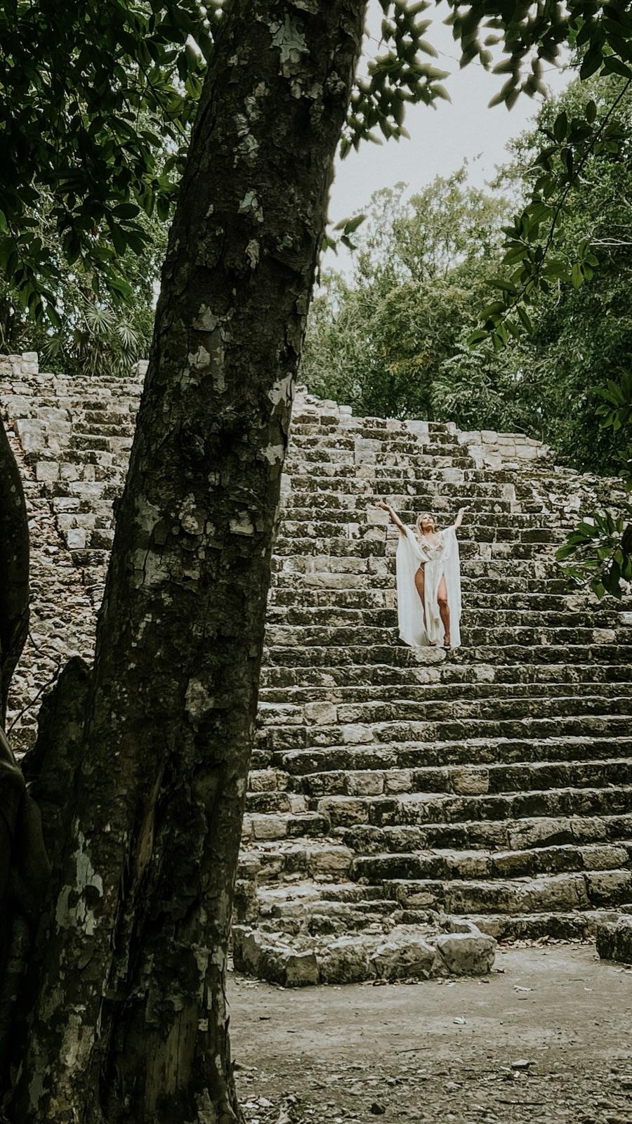 Una persona vestida de blanco de pie con los brazos abiertos en los escalones de piedra de una pirámide maya en Cobá, enmarcada por la selva.