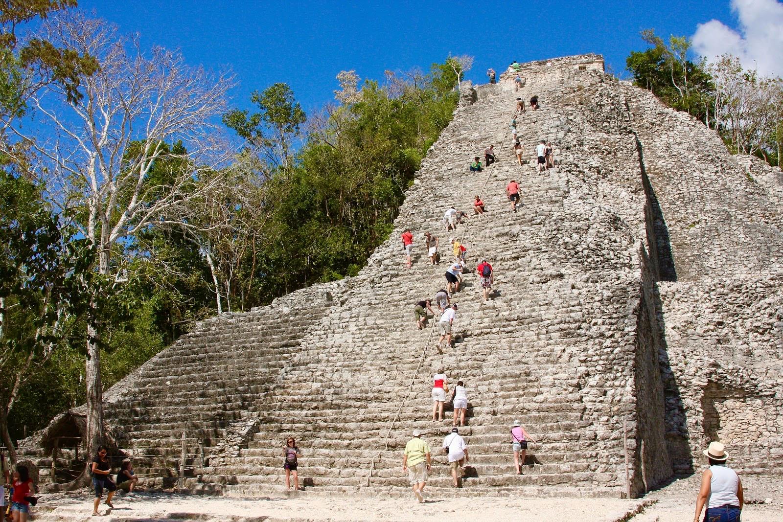 The tall stone Nohoch Mul pyramid at Cobá rising above the jungle, with visitors climbing its steep central staircase under a blue sky.