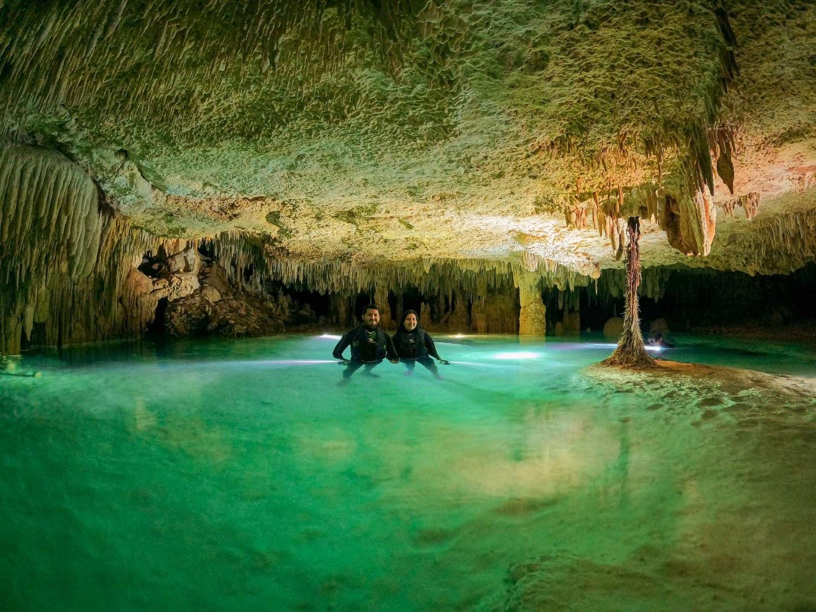 Two people wade through the clear turquoise water of an underground cavern, with stalactites and rock formations reaching down toward the surface.