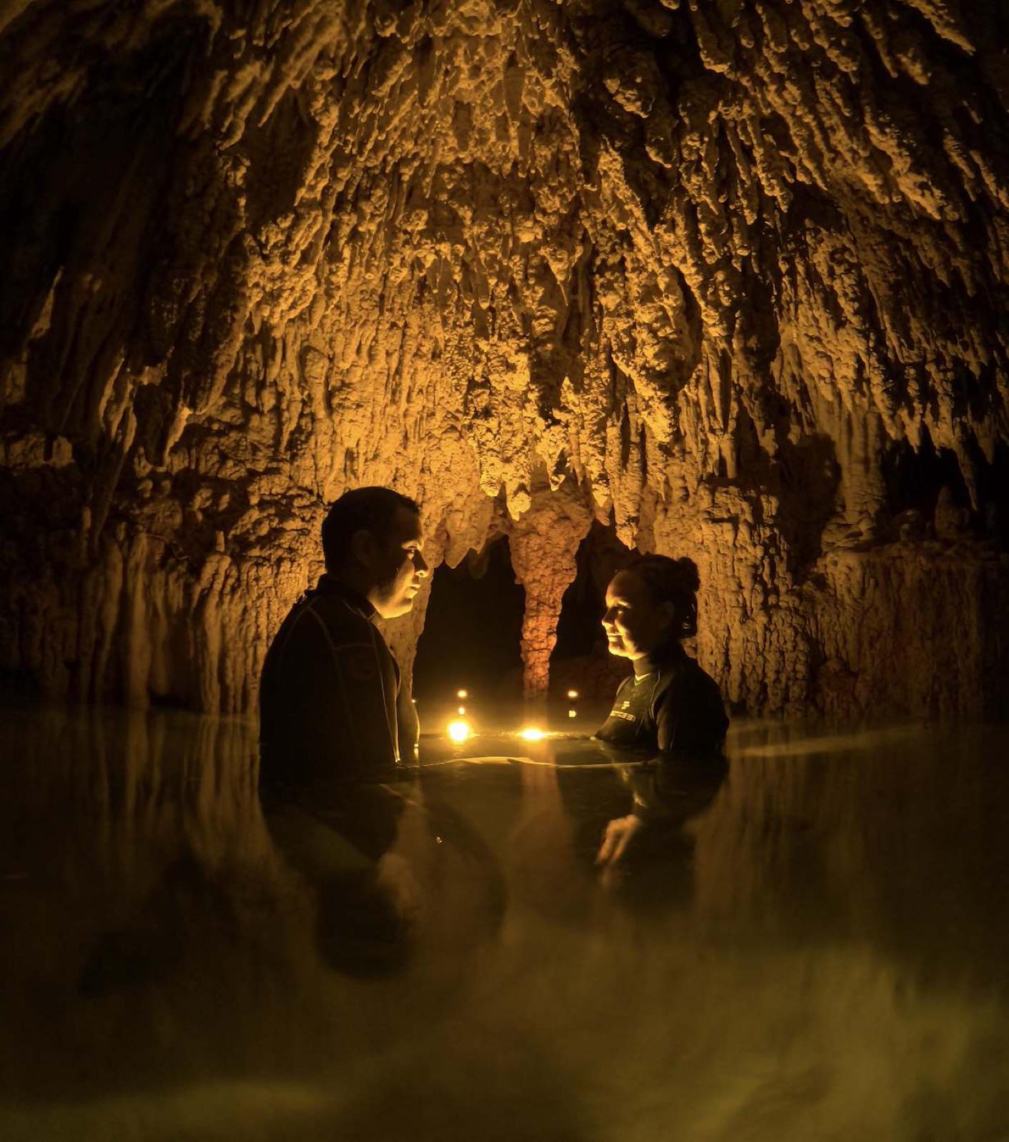 A couple stands facing each other in the dark still water of a cave, their faces lit by the warm glow of a lantern floating between them.