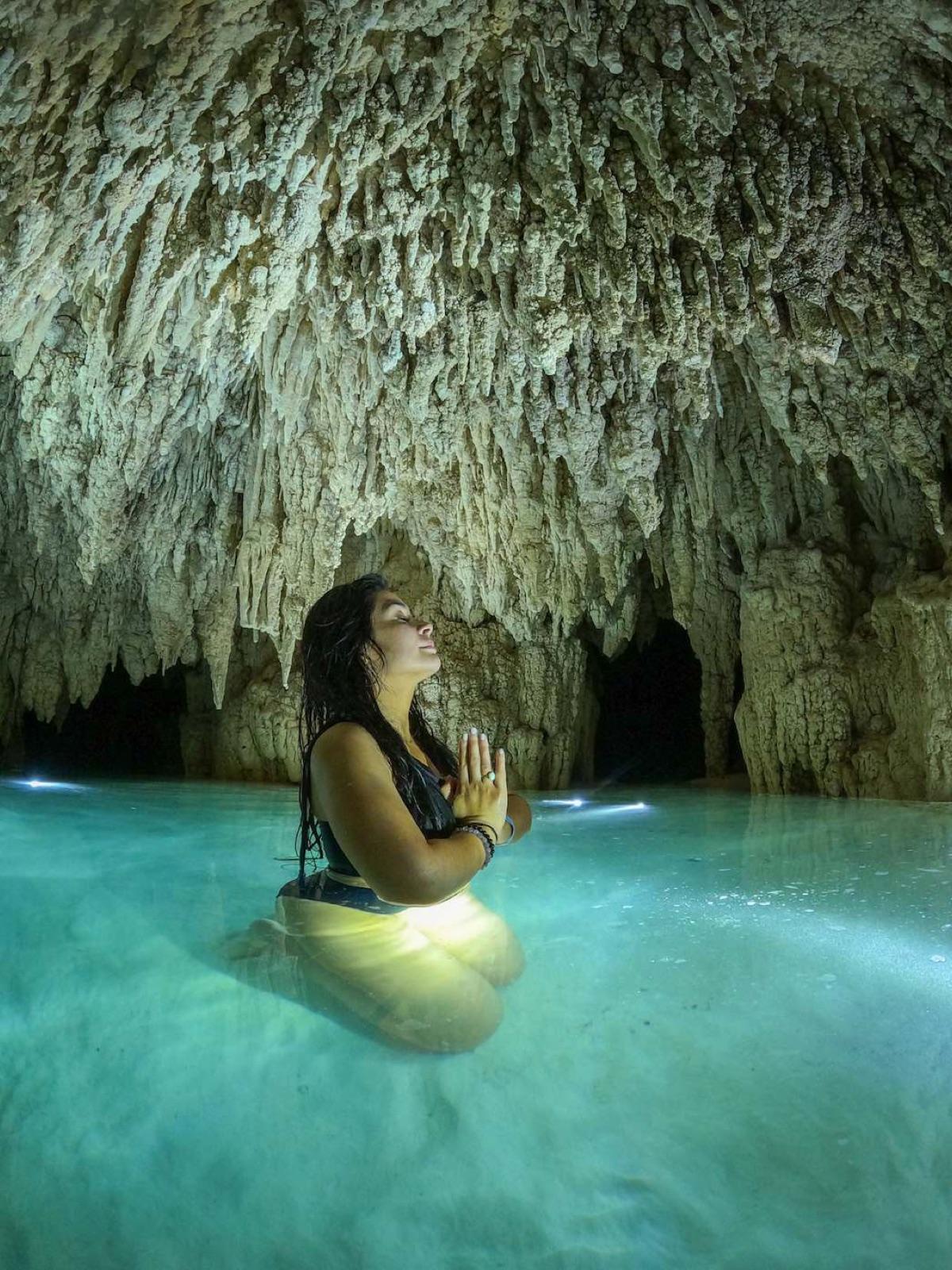 A woman stands in turquoise cave water with hands pressed together in a meditative gesture, a lantern glowing beside her beneath a ceiling of stalactites.