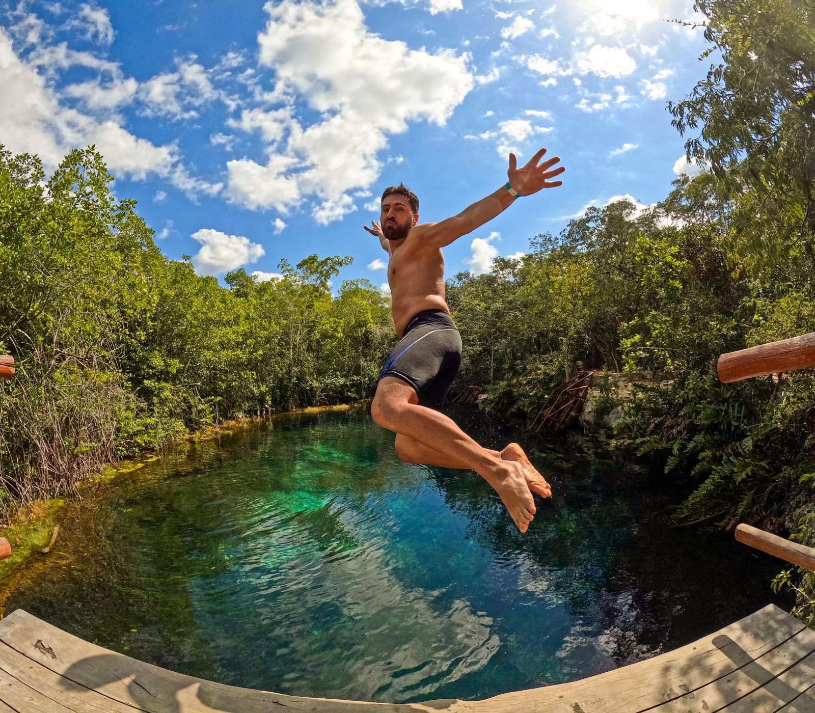 A man leaps arms-open into a turquoise open-air cenote ringed by jungle, seen from a wooden platform.