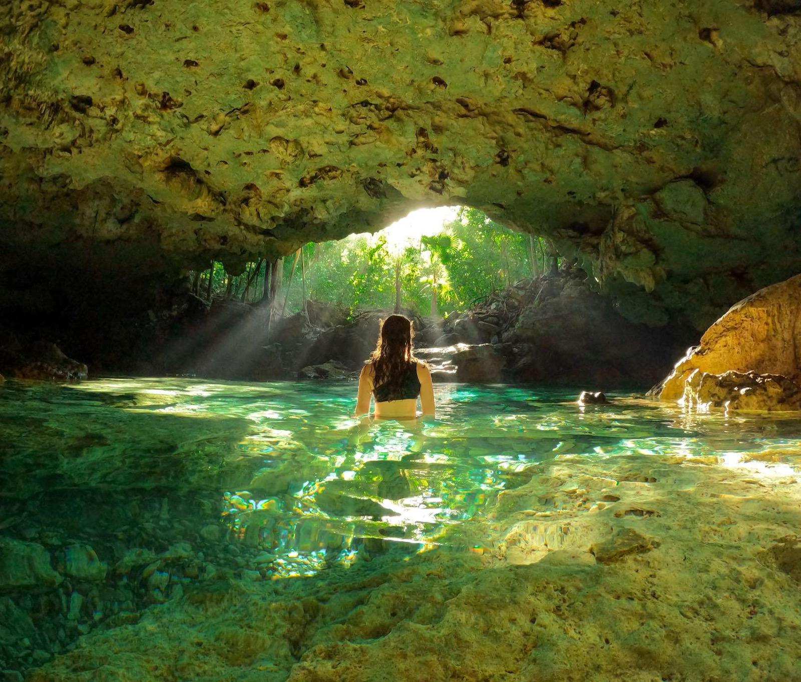 A woman wades into a cavern cenote as a shaft of sunlight breaks through the opening onto clear turquoise water.