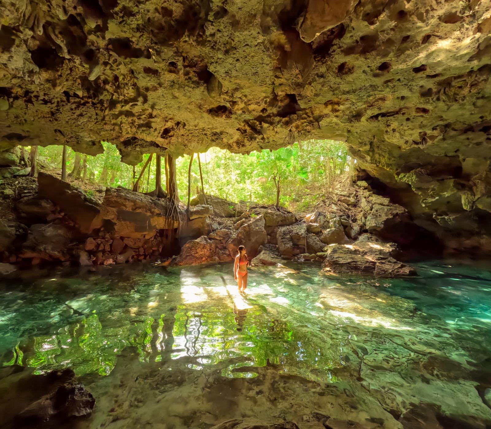 A lone swimmer stands in a cavern cenote as daylight streams in through the jungle-framed entrance.