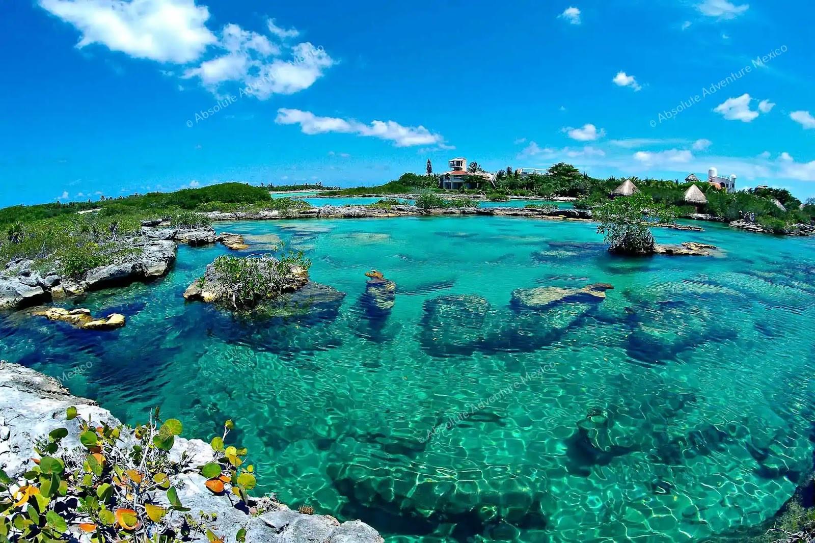 Aerial view of the Yal-Kú lagoon: clear turquoise water, rocky coves and jungle under a bright blue sky.