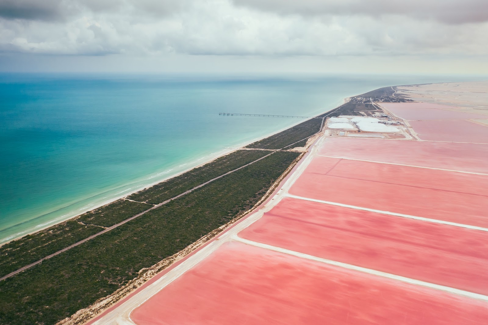 Aerial view of the pink lagoons of Las Coloradas meeting a green shoreline strip and the turquoise sea under a cloudy sky
