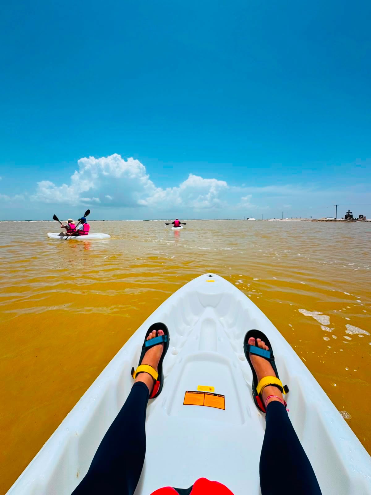 View from the bow of a kayak on an evaporation pond at Las Coloradas, with two other kayaks paddling ahead under a blue sky