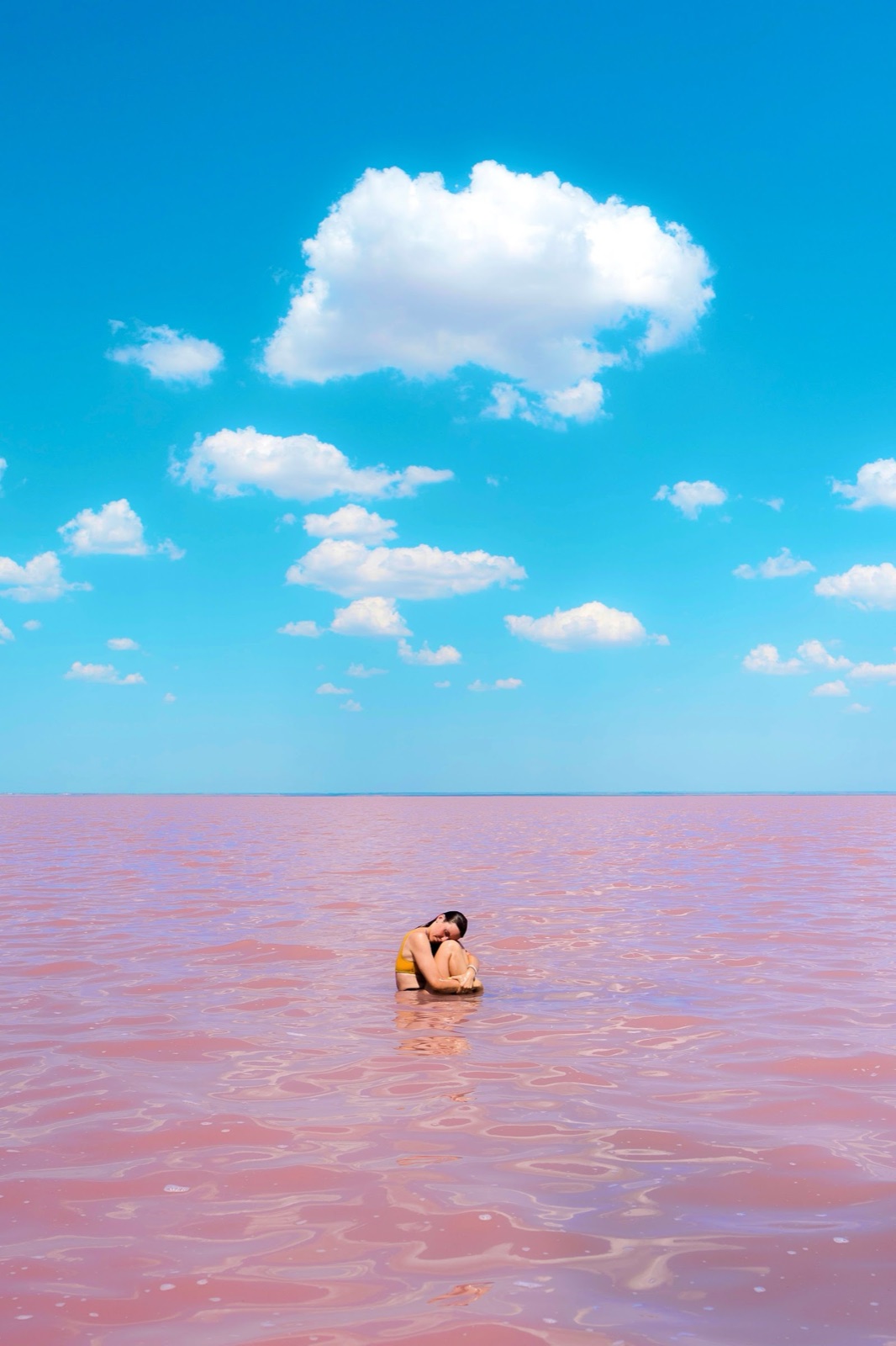 A person sitting in the shallow pink lagoon water beneath a bright blue sky dotted with white clouds