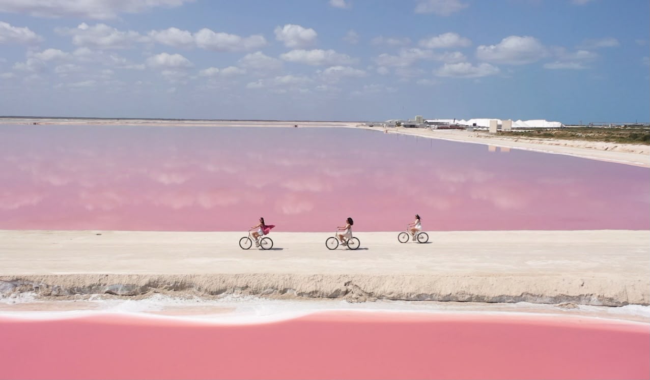 Three cyclists riding along a white salt causeway between two pink lagoons at Las Coloradas, with the saltworks in the distance