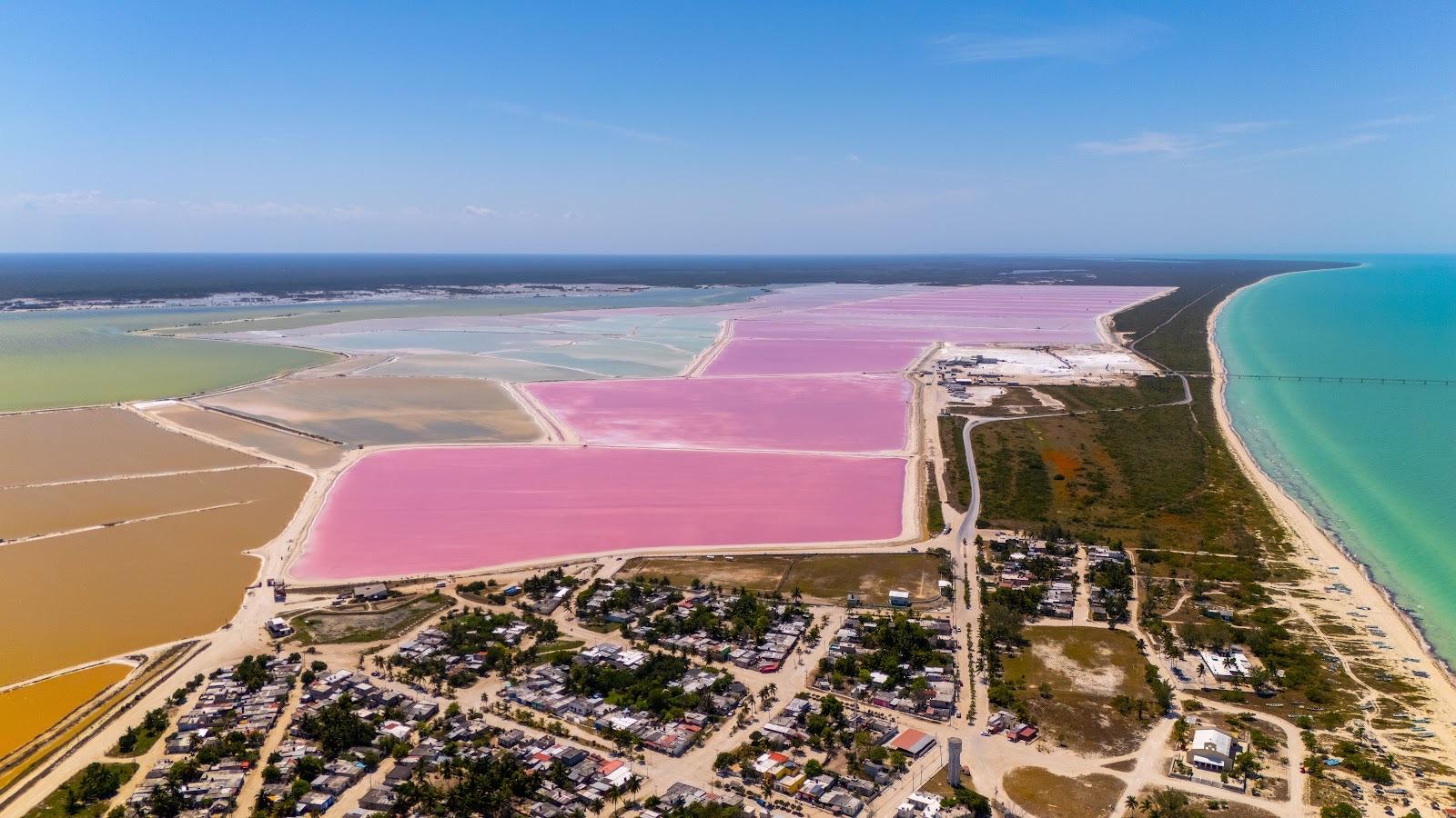 Aerial view of the vivid pink salt lagoons of Las Coloradas beside a coastal town and the turquoise Gulf under a clear blue sky