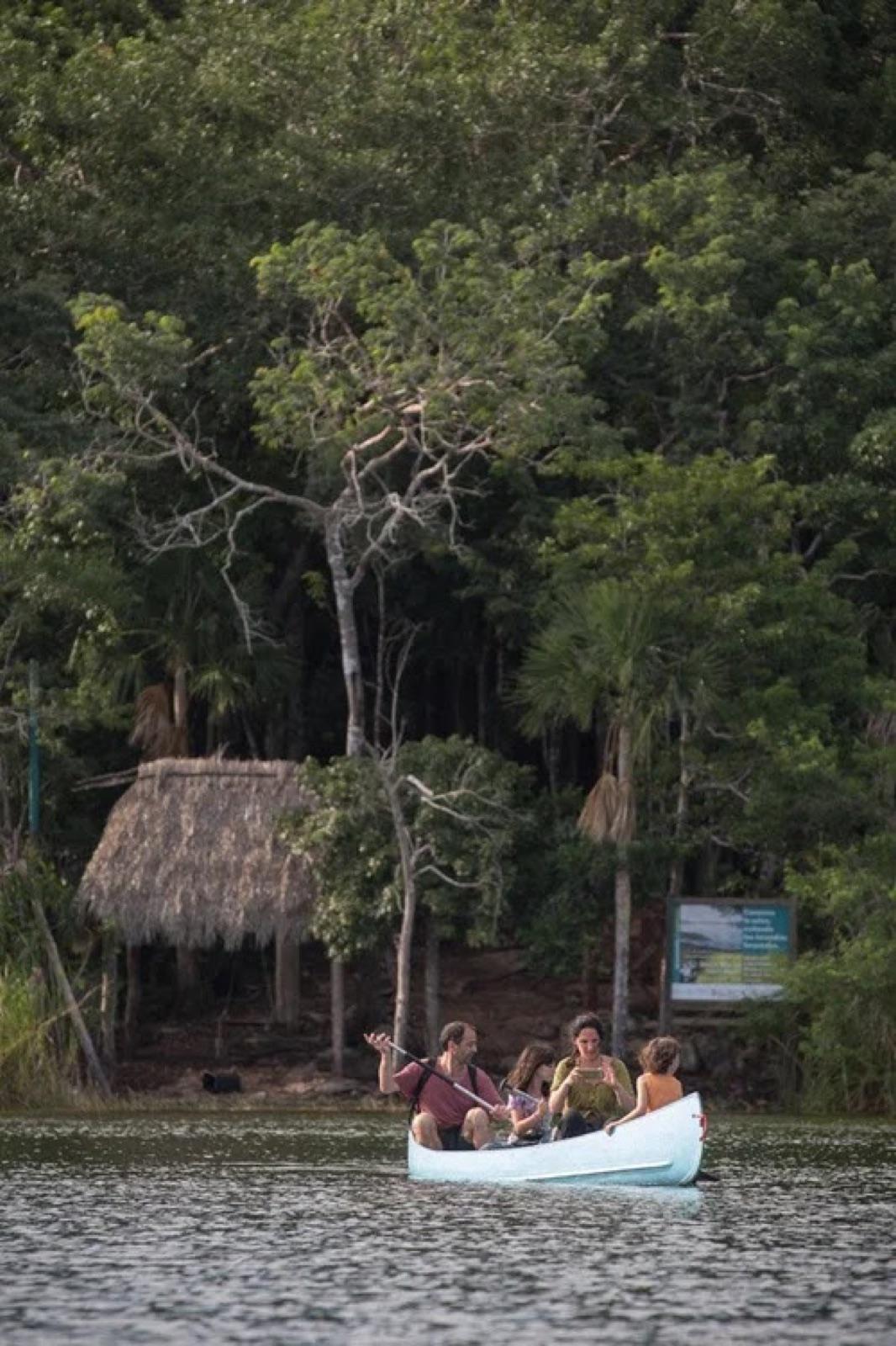 Visitantes reman en canoa por la laguna frente a una palapa en la orilla de la selva.