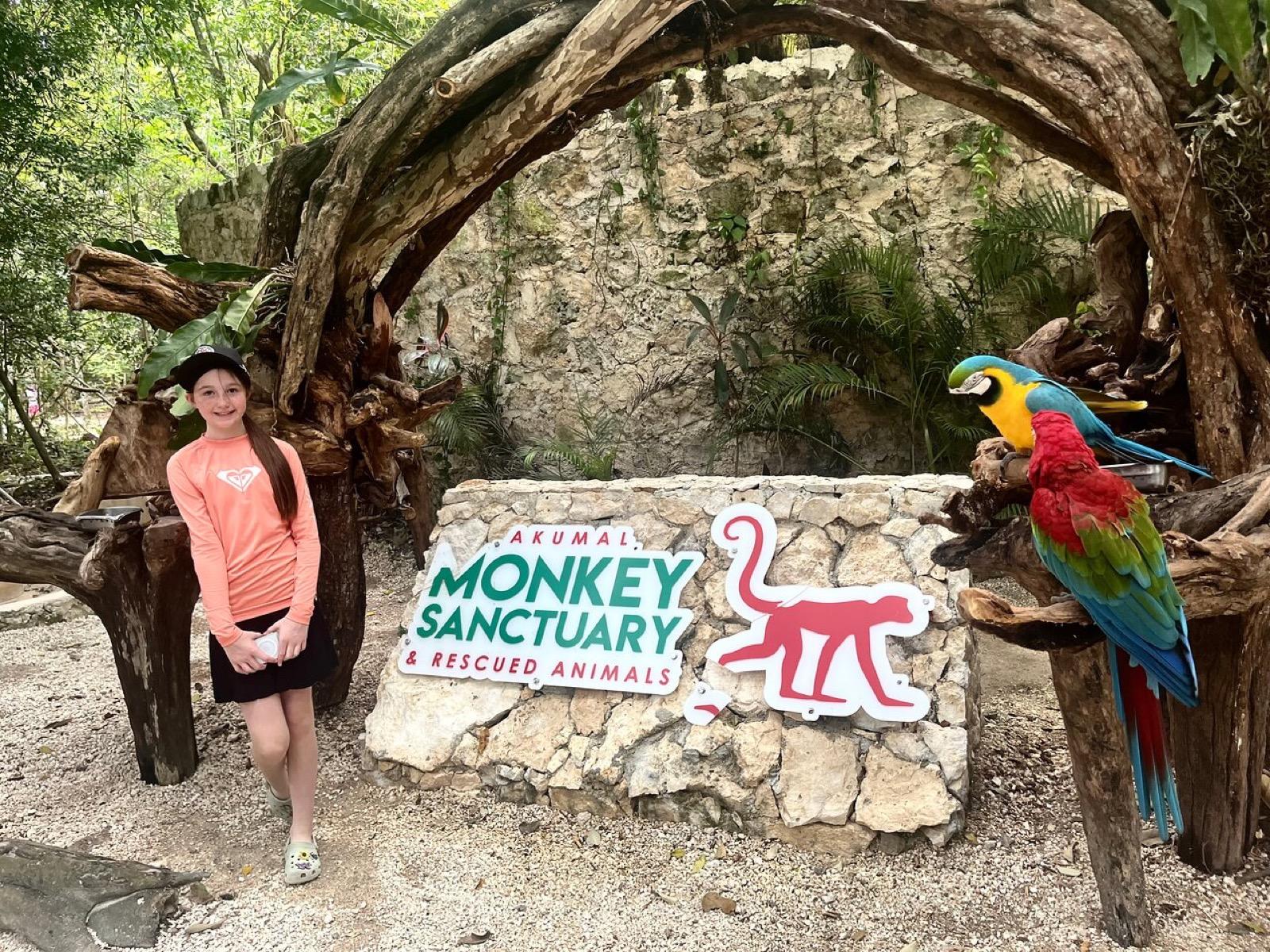 A visitor at the entrance of the Monkey Sanctuary beside its stone sign and resident macaws.