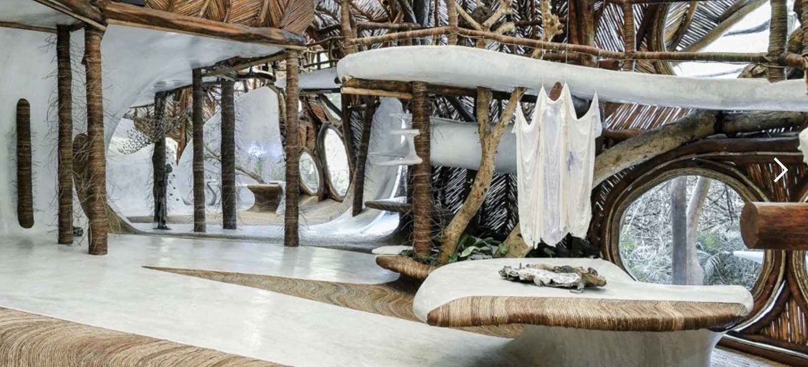 Interior of SFER IK with tree-trunk columns, a woven bejuco ceiling, round windows opening to the jungle and a white garment hanging in the space.
