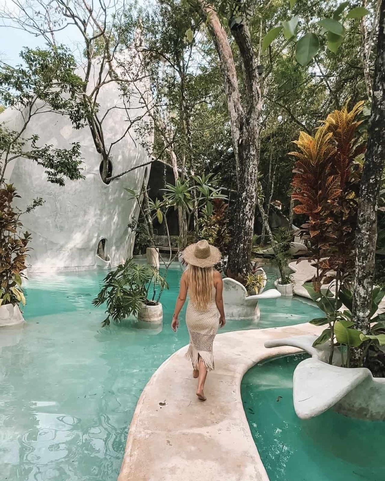 Grounds of the Azulik Uh May / SFER IK complex: a woman in a sun hat walks a curved stone path between turquoise pools in the jungle, with organic white architecture behind.