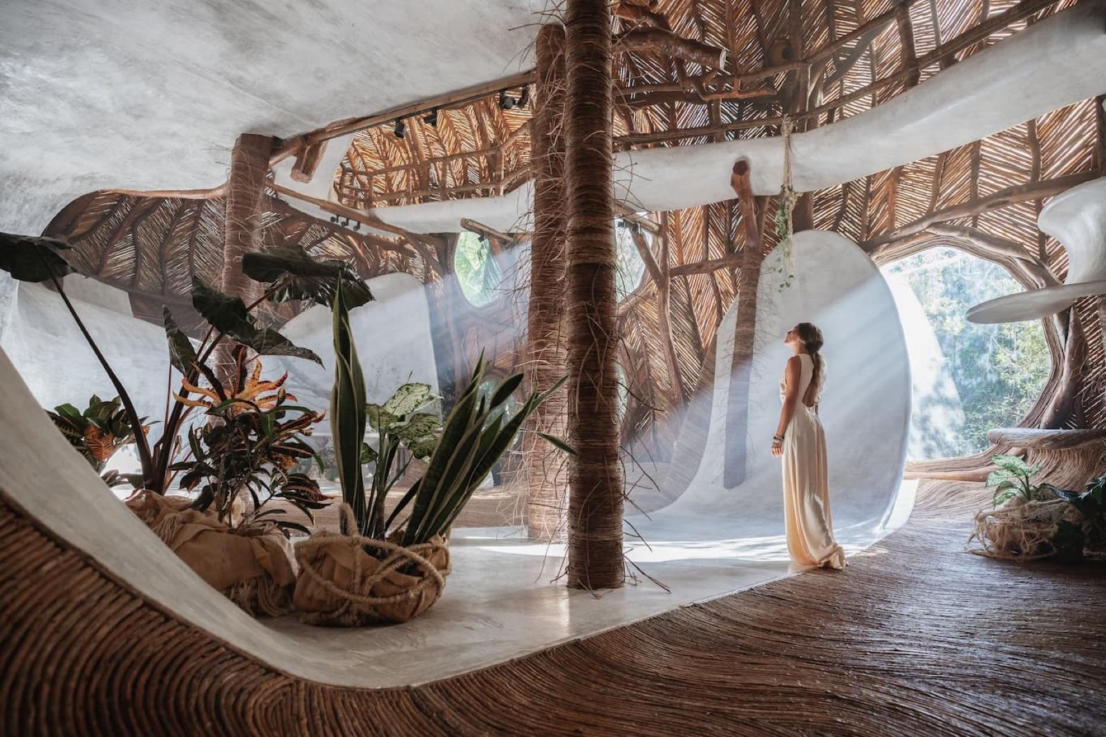 Interior of SFER IK Museion in Tulum: a woman in a flowing white dress stands in a sunlit gallery with a woven bejuco ceiling, curved white organic walls, a polished wood floor and tropical plants.