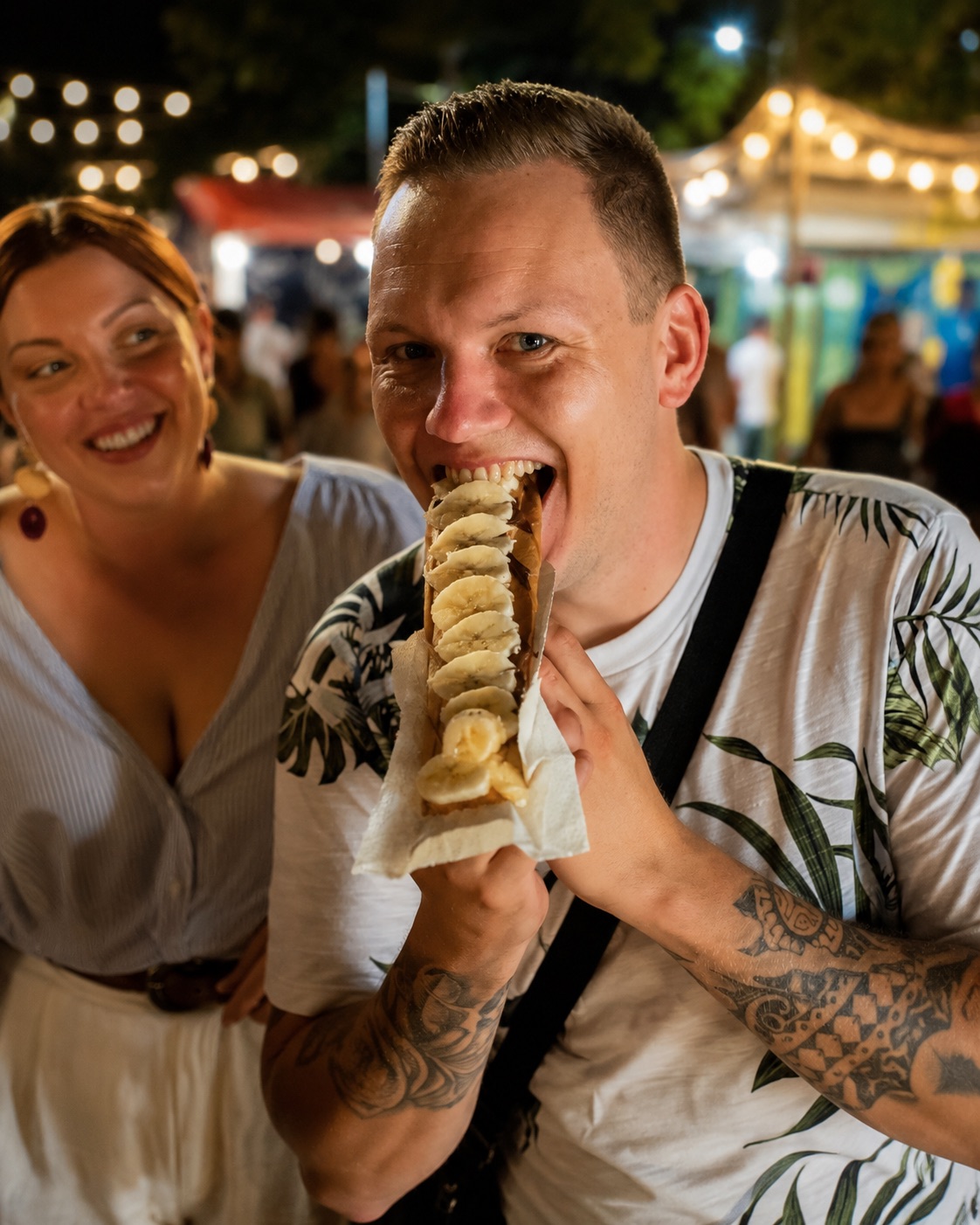 A couple at a night market, one biting into a fresh street-food snack under warm string lights.