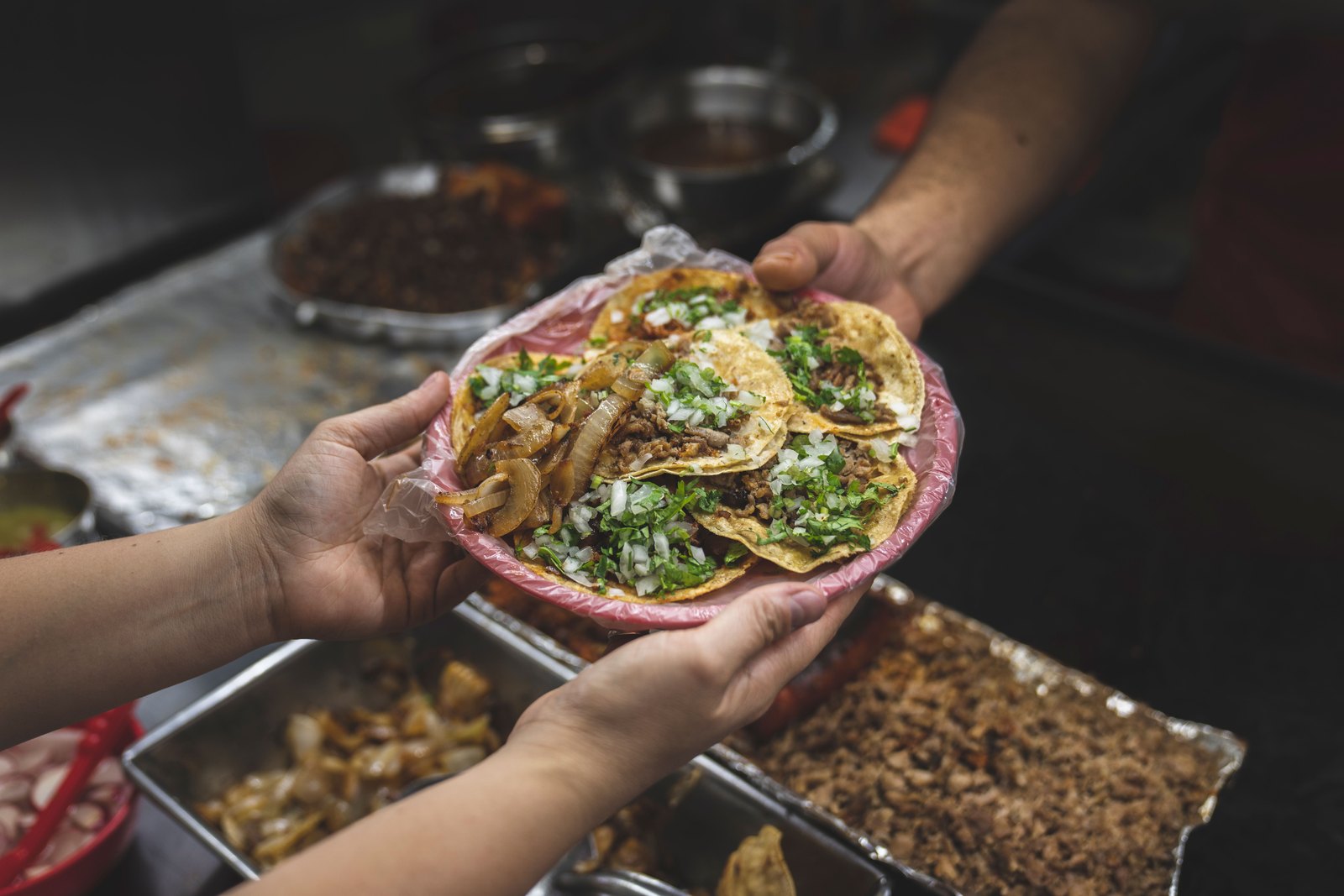 A cook handing a plate of tacos across the counter, trays of stewed fillings lined up behind him.