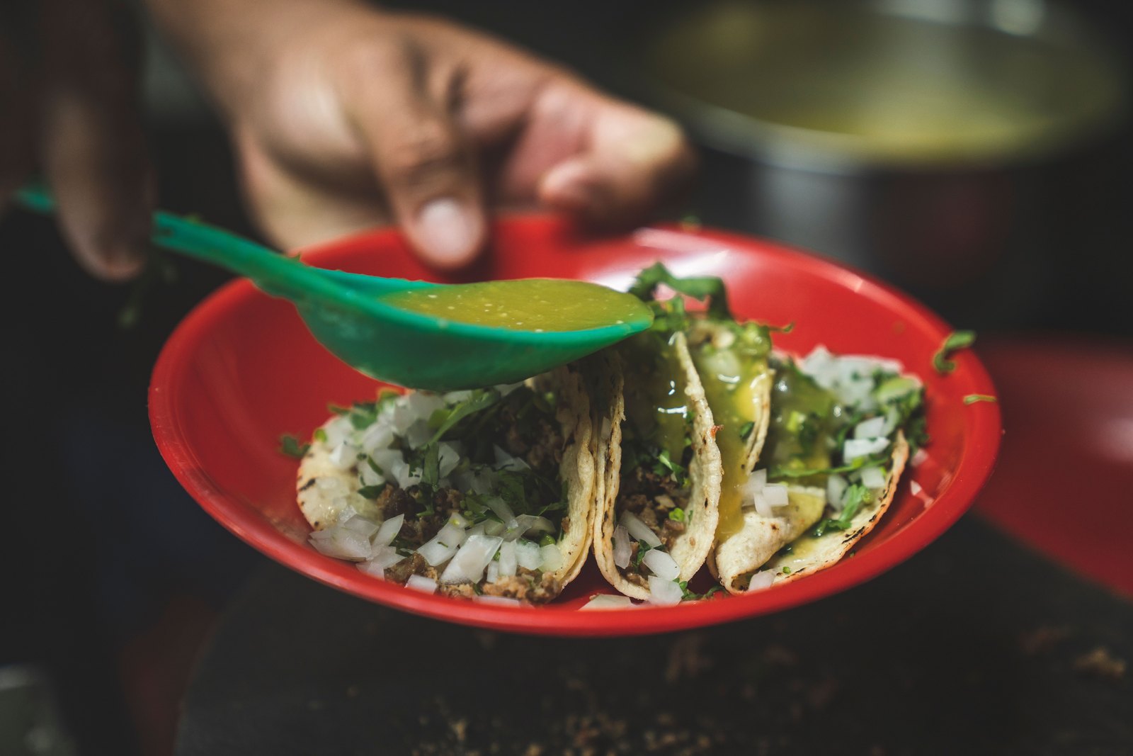 Green salsa spooned over three tacos topped with white onion and coriander on a red plate.