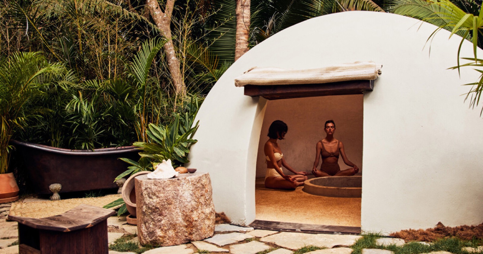 Dos personas meditan en la entrada en arco de un temazcal de domo blanco, enmarcadas por plantas tropicales.