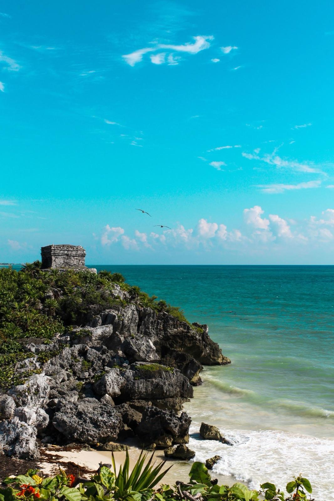 A small Maya temple perched on the rocky Tulum cliff above the turquoise Caribbean Sea, birds gliding across a blue sky