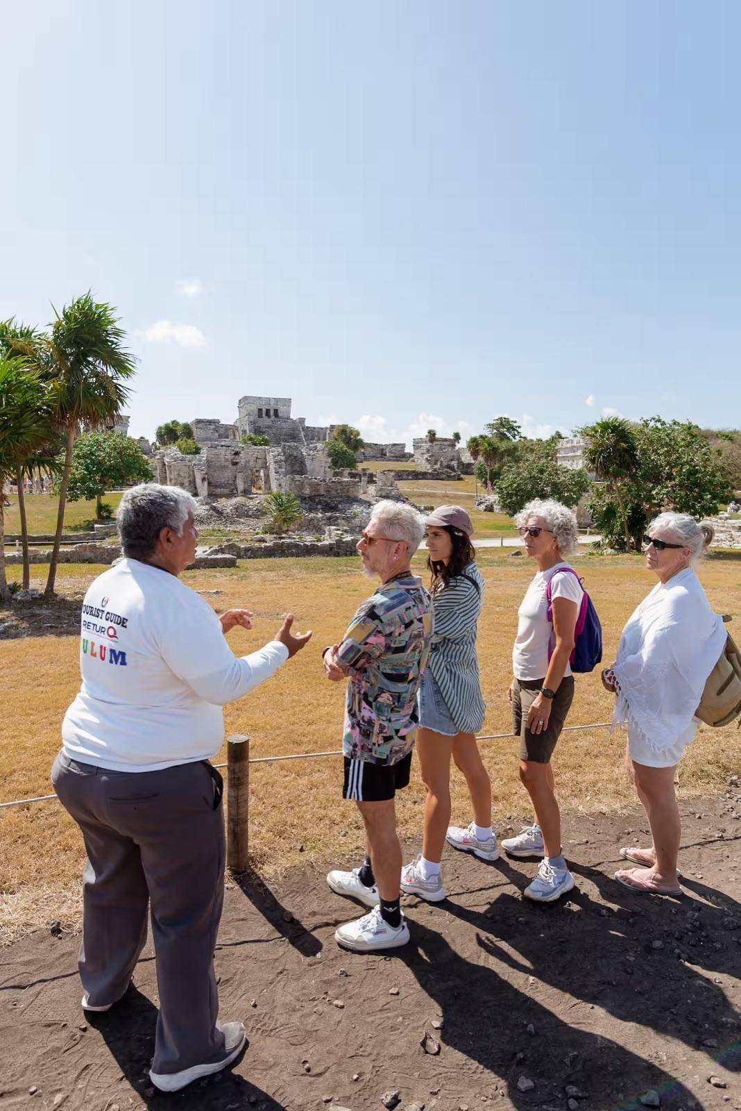 A local guide leads a small group of guests at the Tulum ruins, the main stone pyramid and palm trees behind them on a sunny day