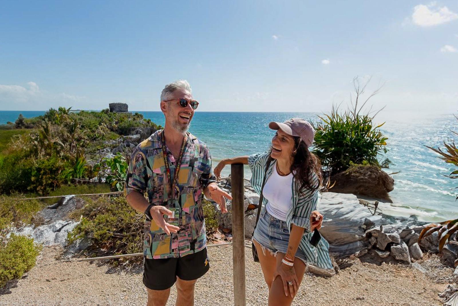 A smiling couple pauses at a cliffside overlook in Tulum, a stone Maya ruin and the turquoise Caribbean Sea behind them under a clear blue sky