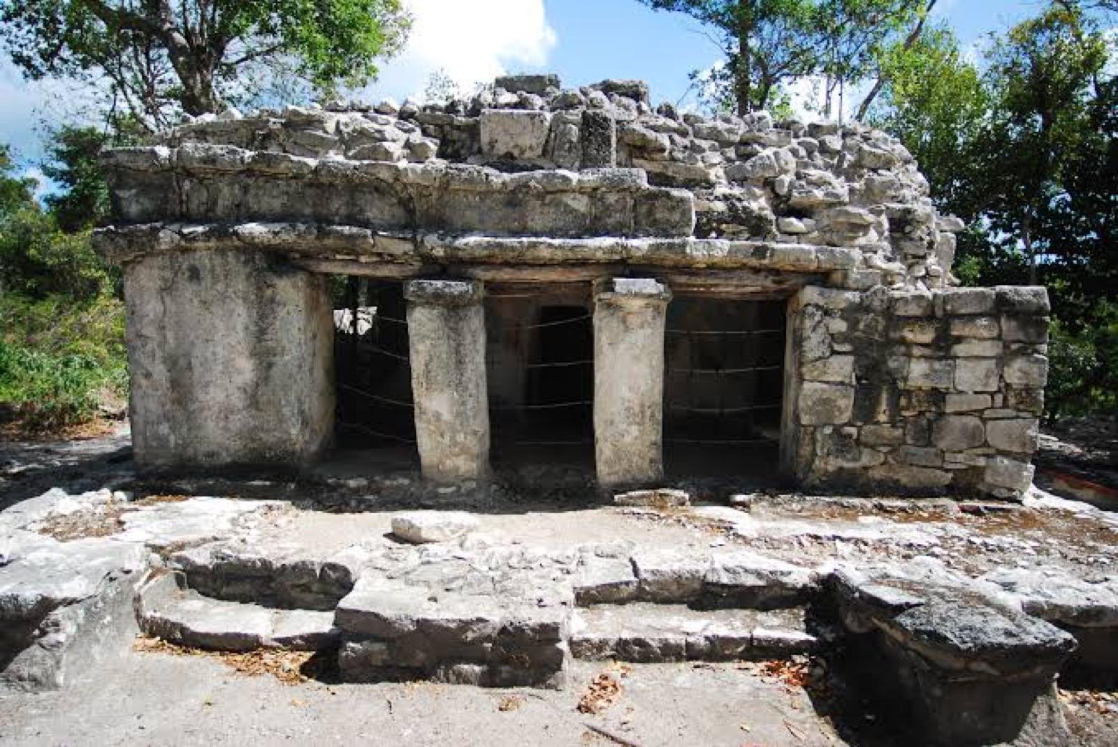 A small Maya stone building with square columns and a doorway in daylight.
