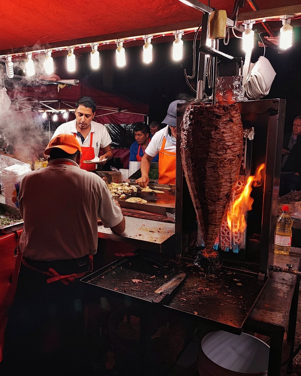 Taqueros at a night street stand, the al pastor spit beside the flame