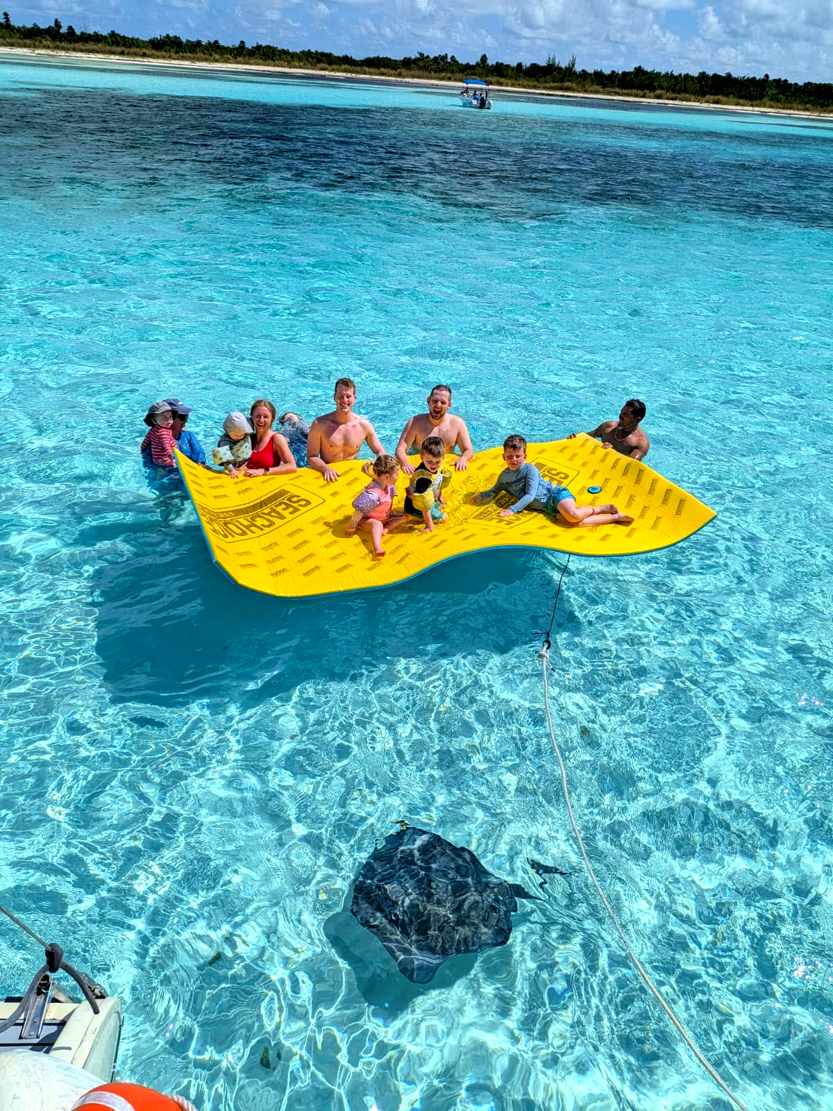 Una familia sobre una alfombra flotante junto al barco, con una raya nadando abajo