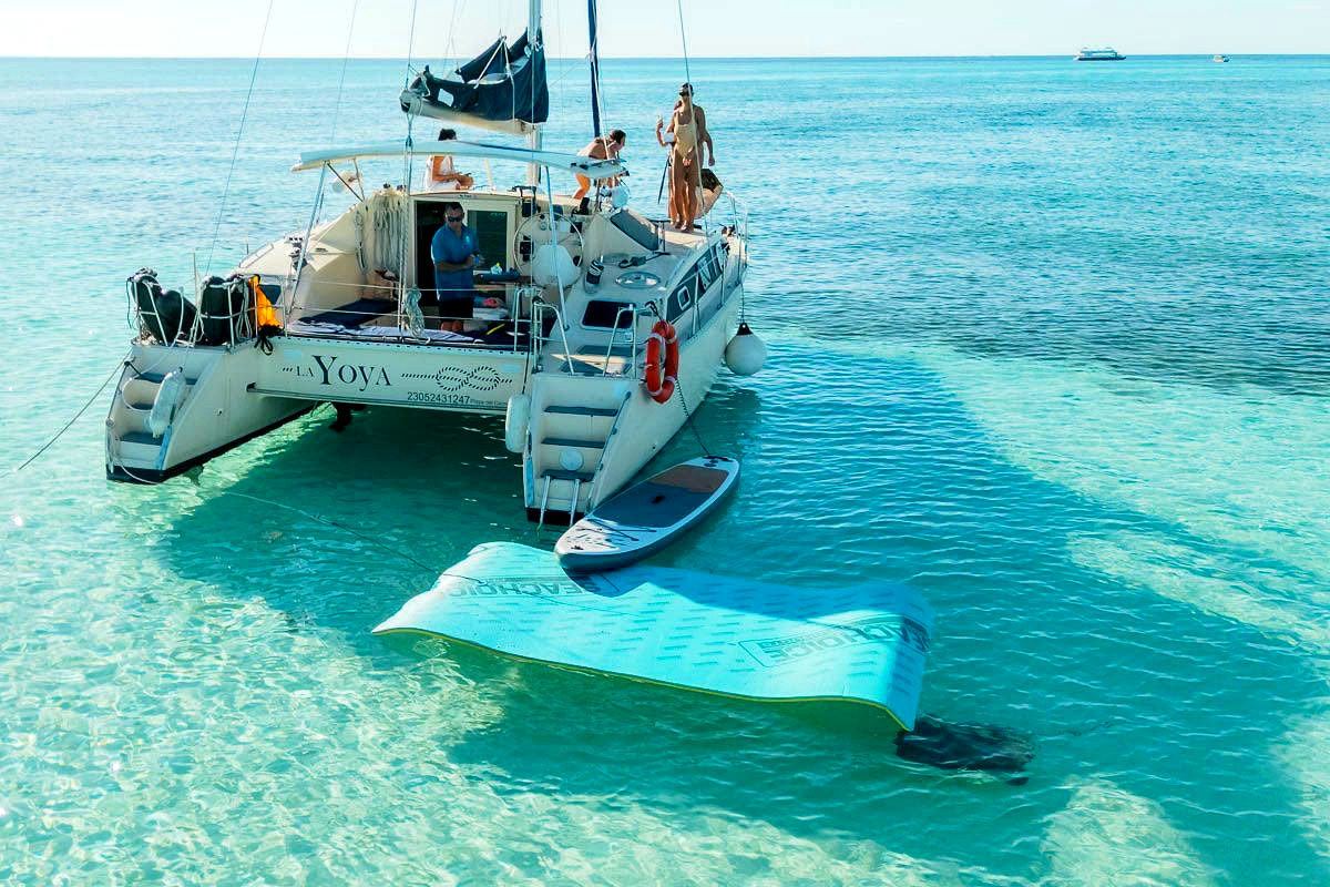 La popa de La Yoya con sus escaleras al agua, un paddle board y una alfombra flotante sobre agua clara