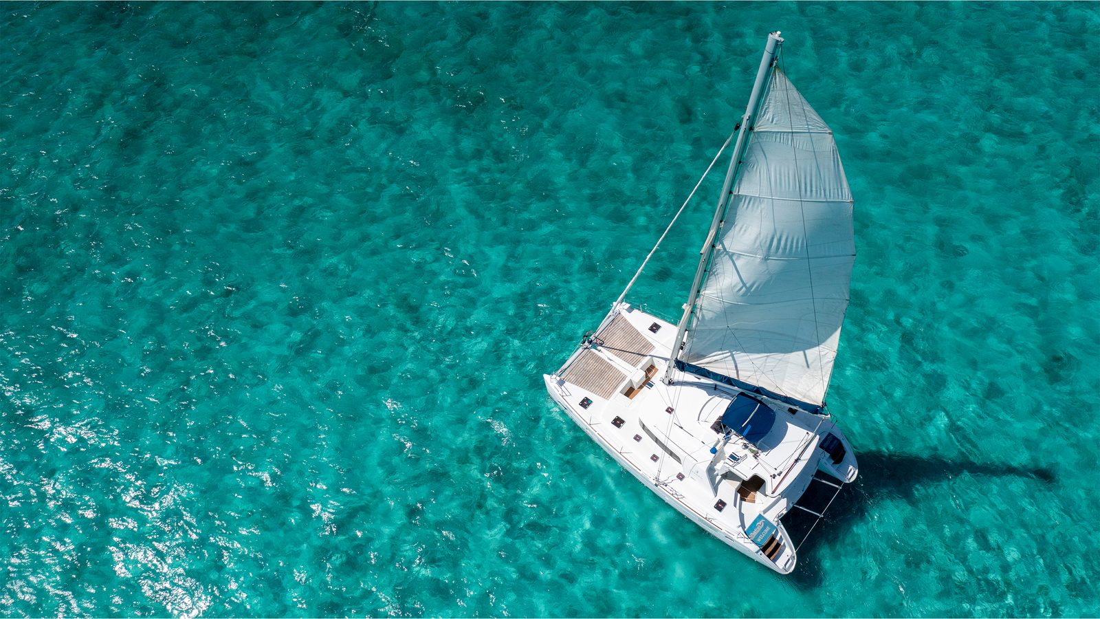 Quidam under sail over clear turquoise water, seen from above
